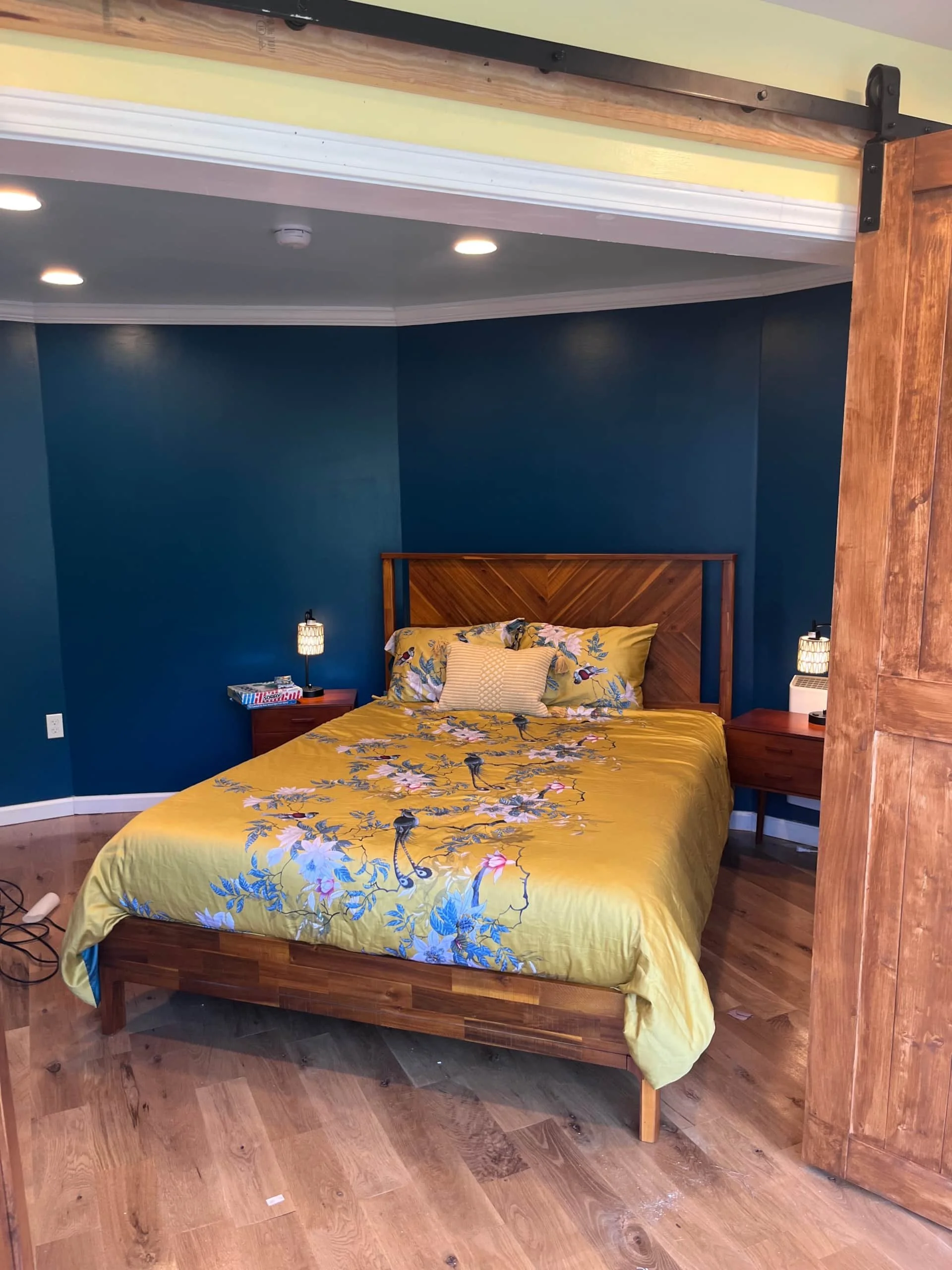 A bedroom with a wooden bed frame, yellow floral bedding, dark blue walls, and two small nightstands with lamps on either side.