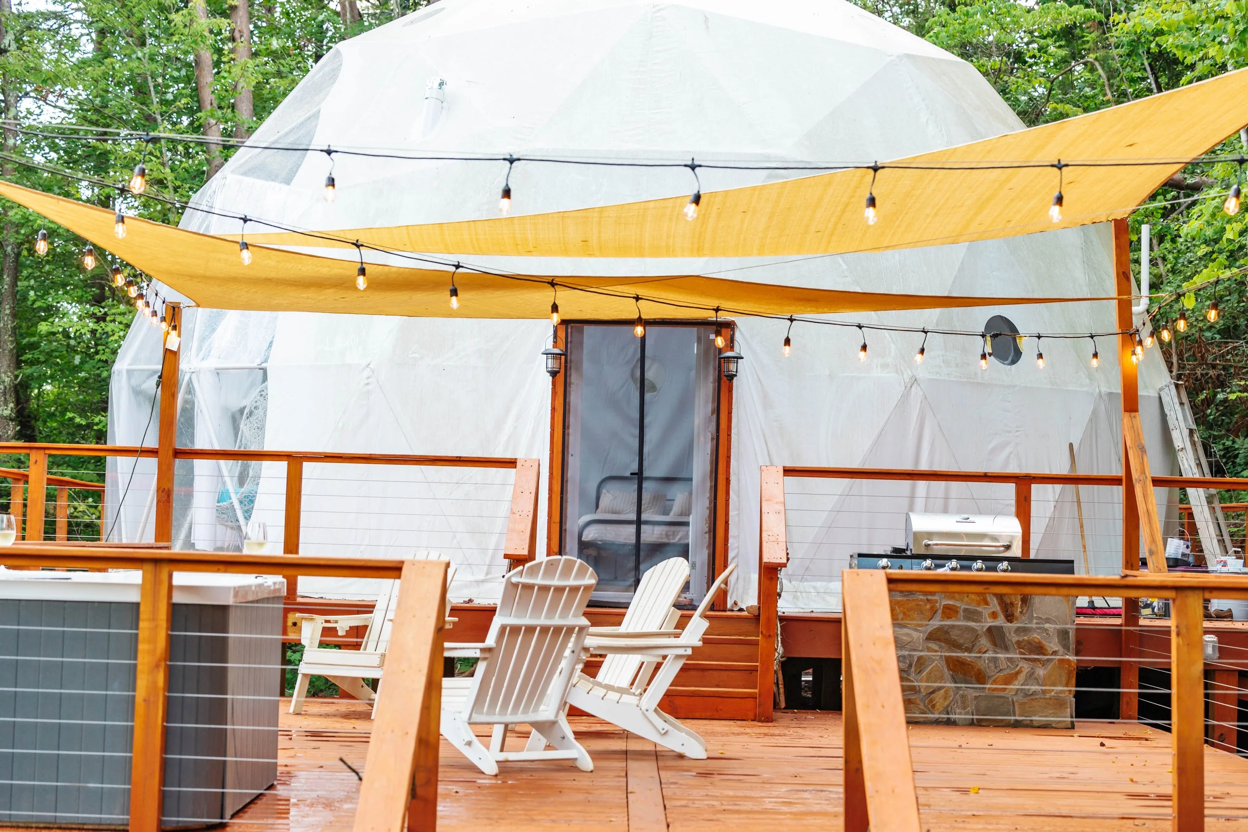 A rustic outdoor deck with Adirondack chairs, string lights, a grill, and a small dome tent in a wooded area with trees and green foliage.
