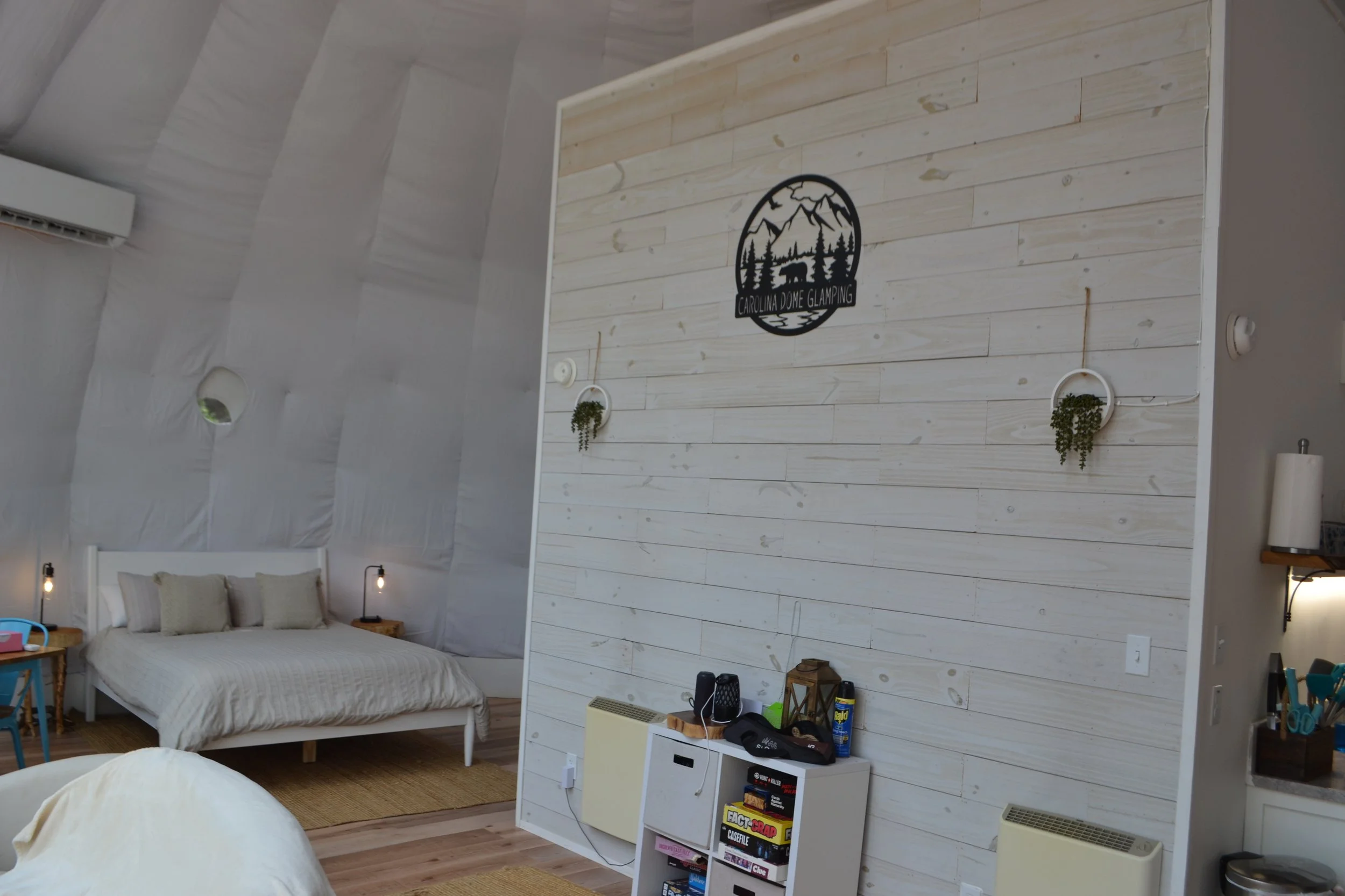 Interior of a cabin with a white bed, nightstands with lamps, a white shiplap feature wall with a Carolina Dome Glamping logo, and a small storage unit with snacks and accessories.