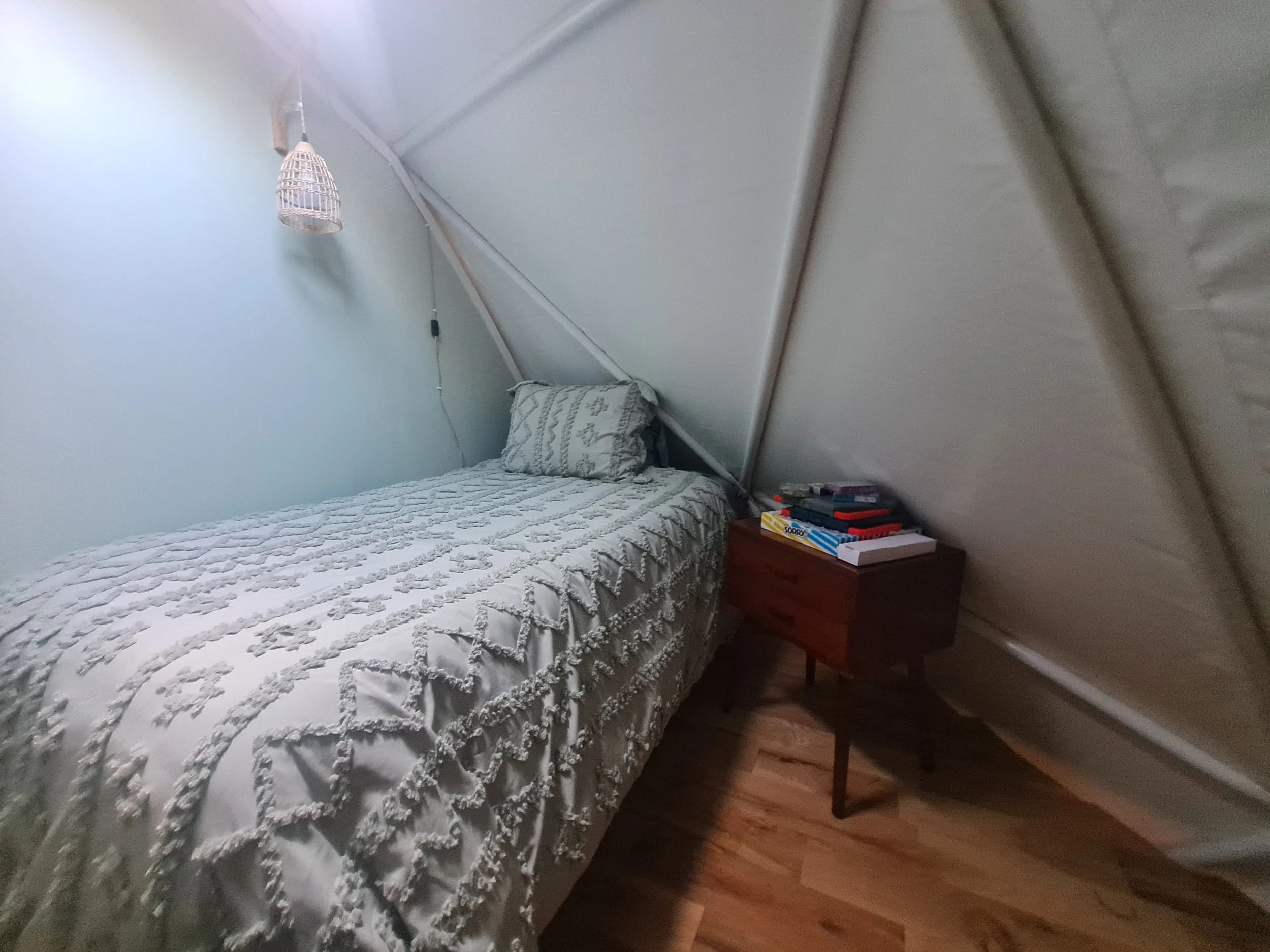 A bedroom with a bed along the left wall, covered with a textured bedspread and pillow. To the right is a wooden nightstand with books and game boxes. The ceiling and wall are sloped, indicating an attic or loft space. A hanging light fixture is abov