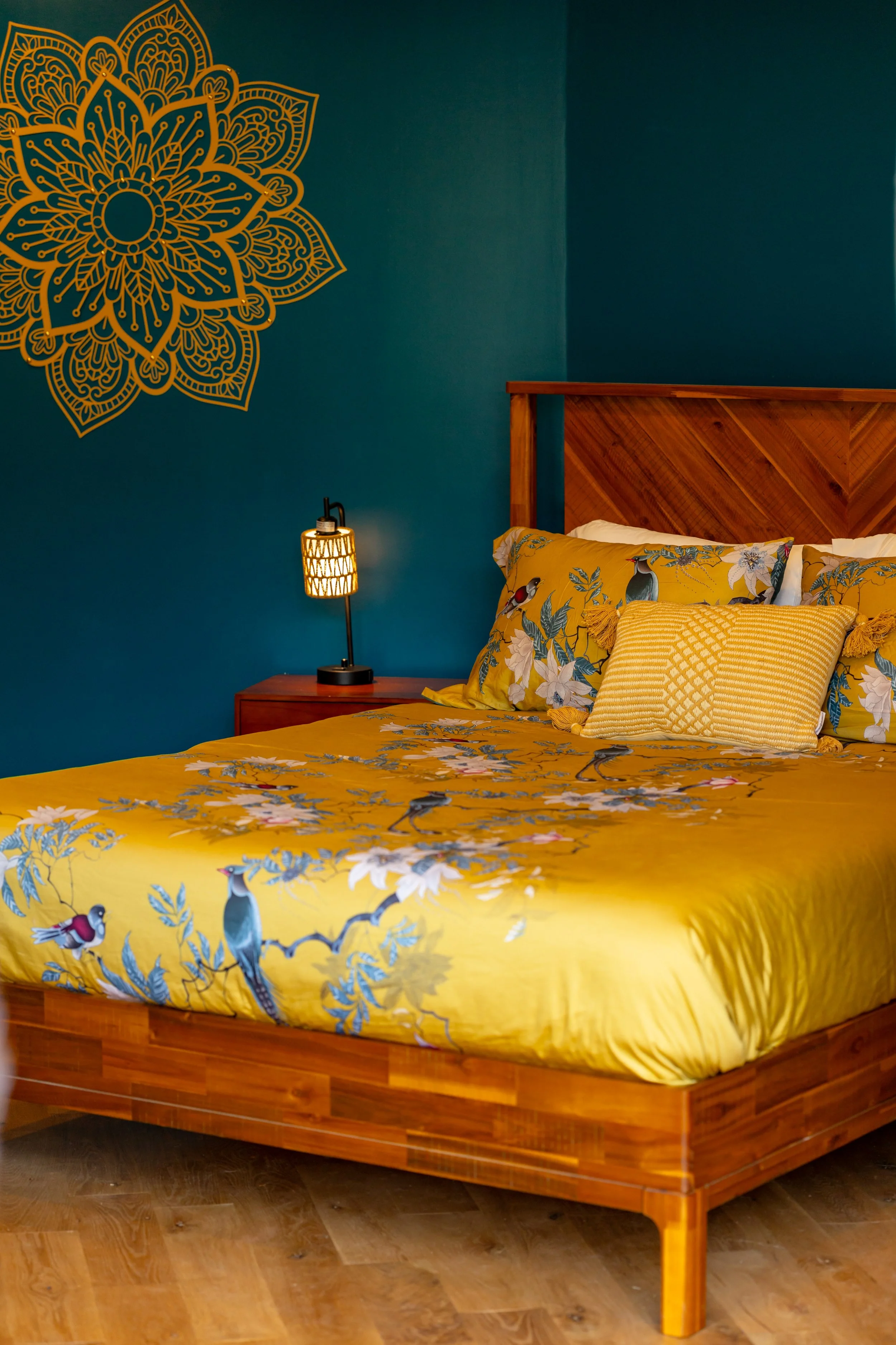 Decorative bedroom with a teal wall featuring a large gold mandala art piece. The bed has a wooden headboard and is dressed in yellow bedding with bird and floral patterns, complemented by matching decorative pillows. A small bedside table with a bla