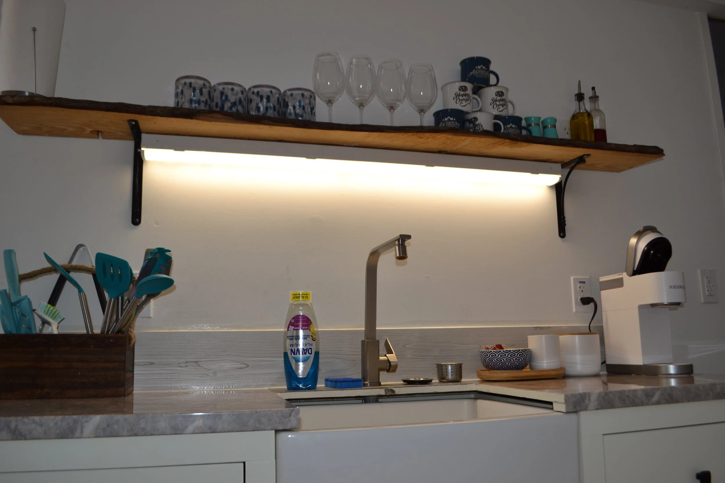 Kitchen sink area with a wooden shelf holding glasses, mugs, and condiment bottles, a dish soap bottle, a utensil holder with cooking tools, and a coffee machine on the counter.