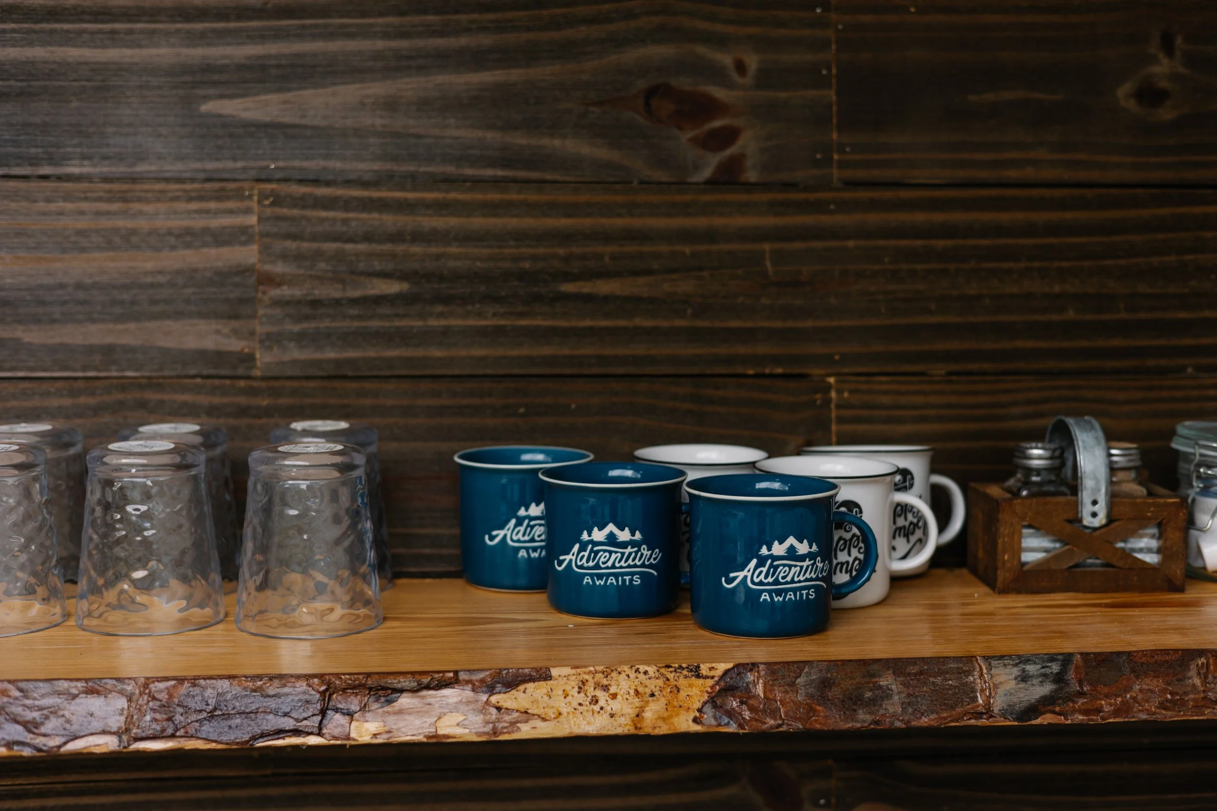 A wooden shelf with upside-down glasses, blue ceramic mugs with white text and mountain logo, white mugs with black text, and a small wooden crate with glass jars inside, all set against a dark wooden wall.