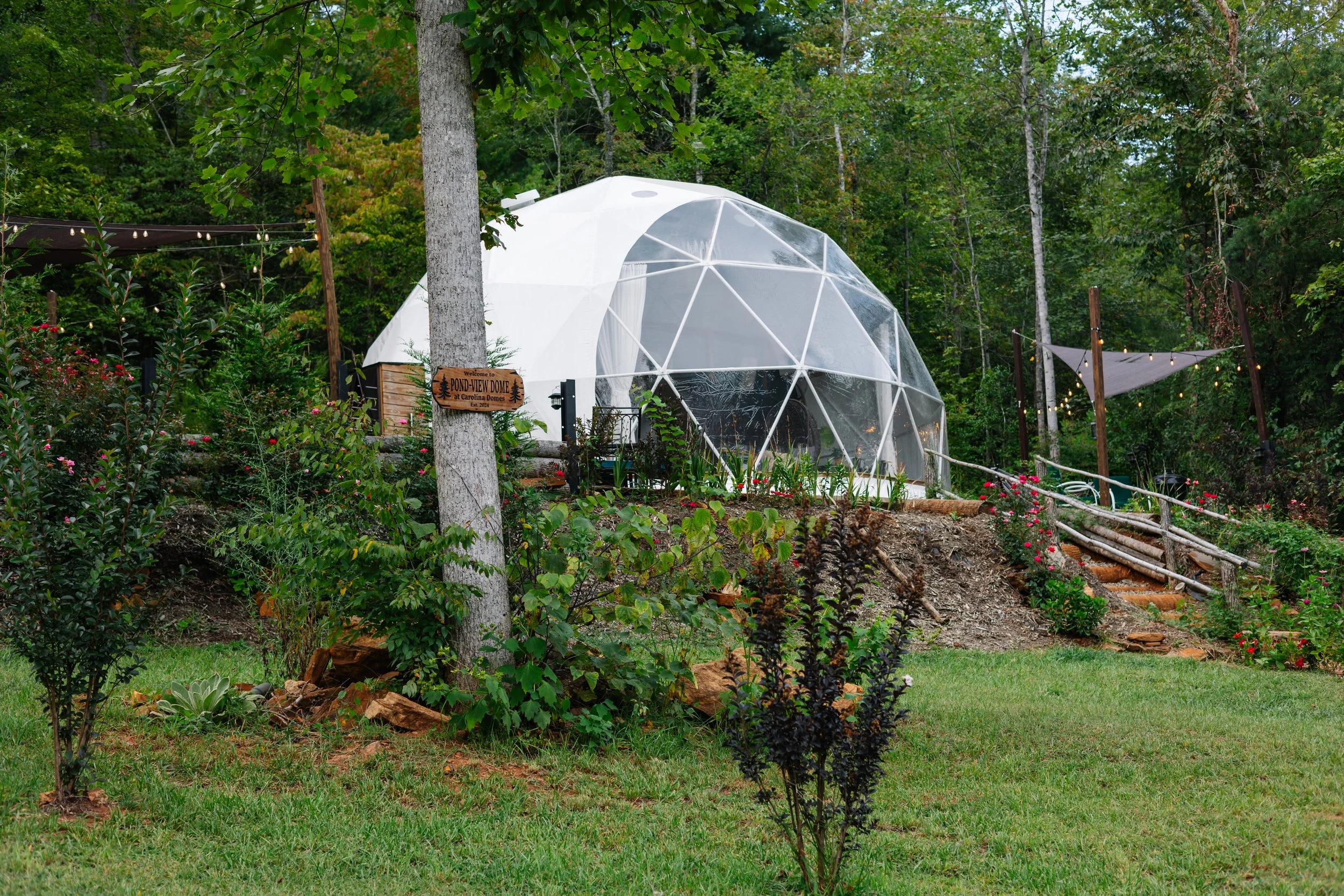 A clear, geodesic dome greenhouse situated on a hillside surrounded by lush greenery and colorful plants and flowers, with a small wooden sign and a garden pathway leading to it.
