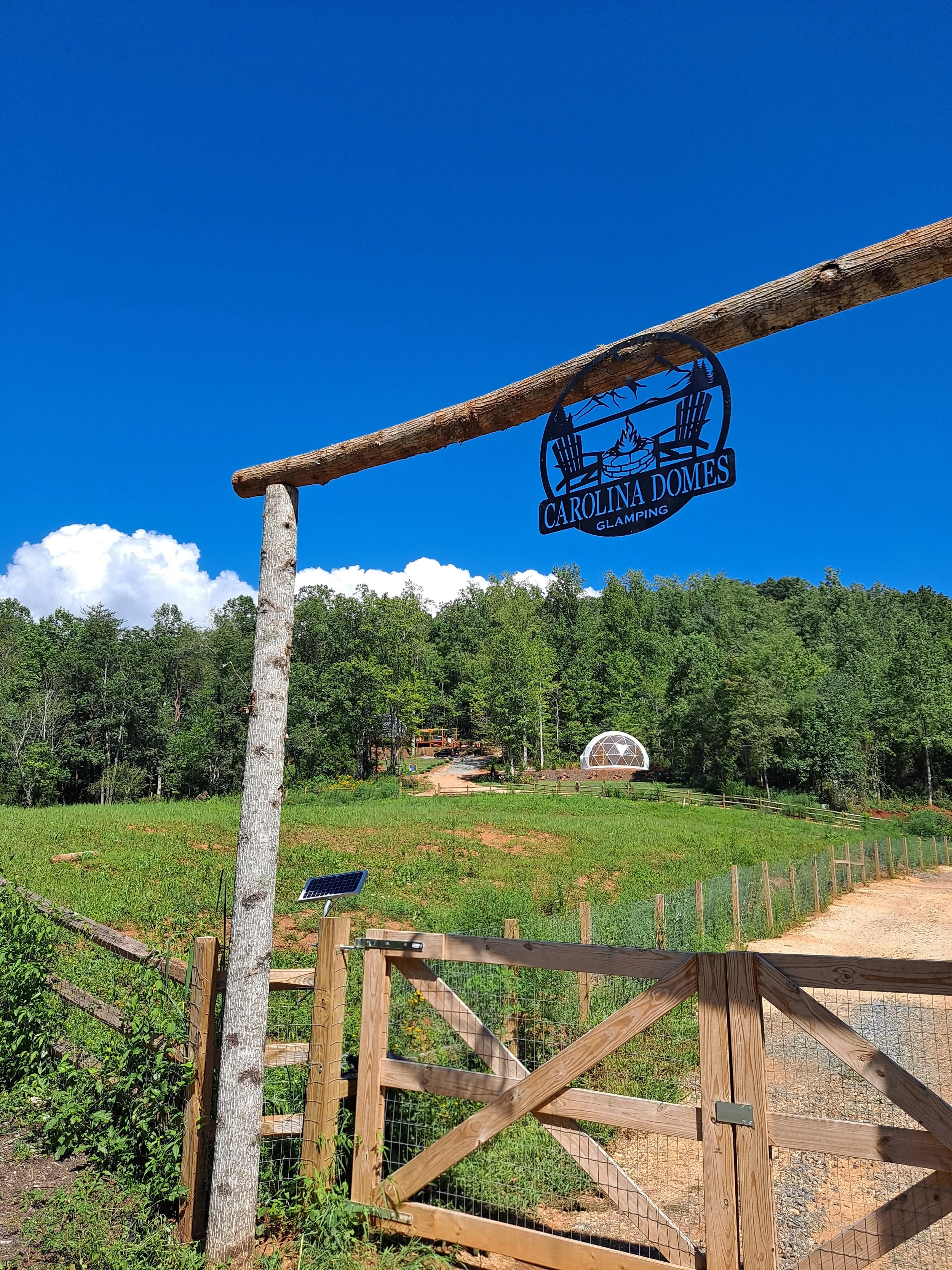 Sign for Carolina Domes Glamping hanging from a wooden beam, with a background of green fields, trees, a geodesic dome, and a clear blue sky.