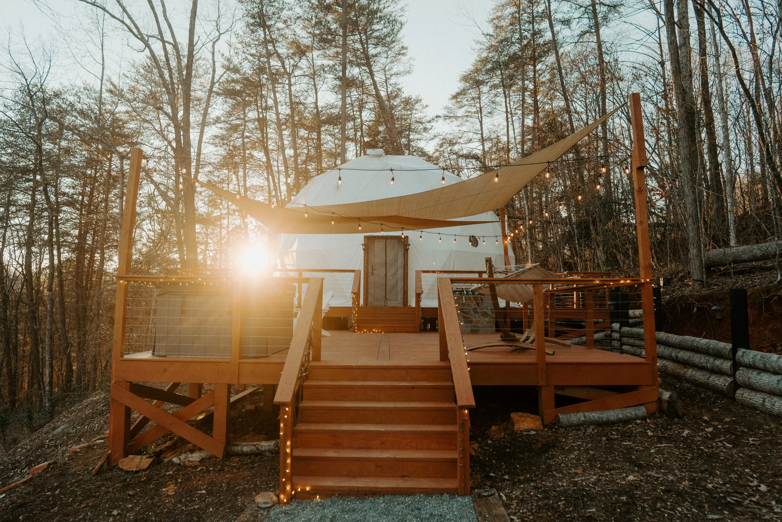 Wooden deck in a forest with string lights, a canopy, and outdoor furniture at sunset.