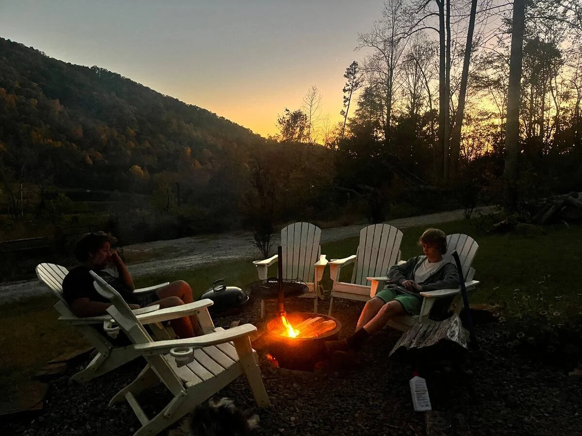 Two children sitting in white Adirondack chairs around a campfire at dusk in a wooded mountain area.