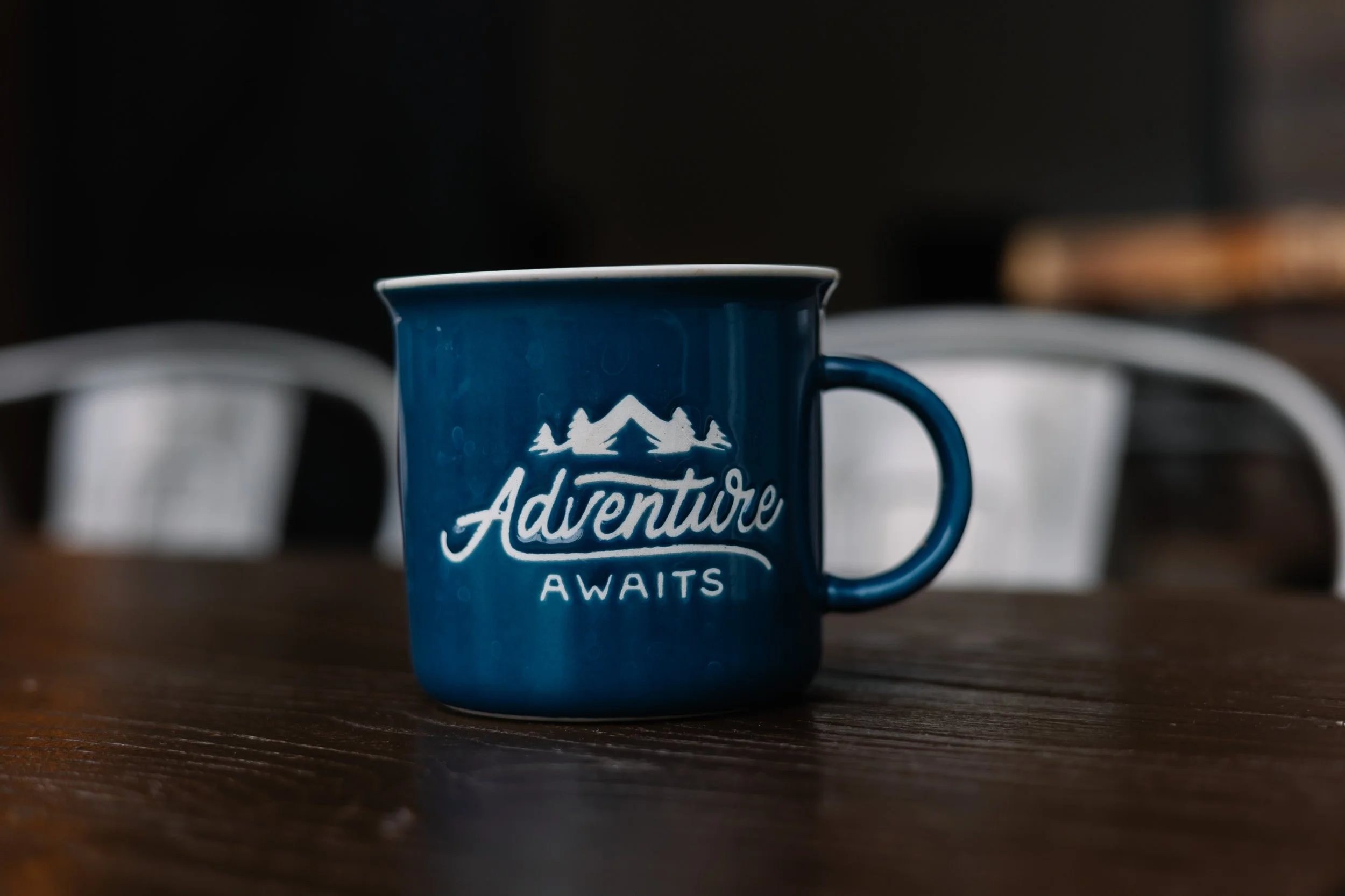 A blue mug with white lettering that says 'Adventure Awaits' and a mountain and trees graphic, placed on a wooden table.