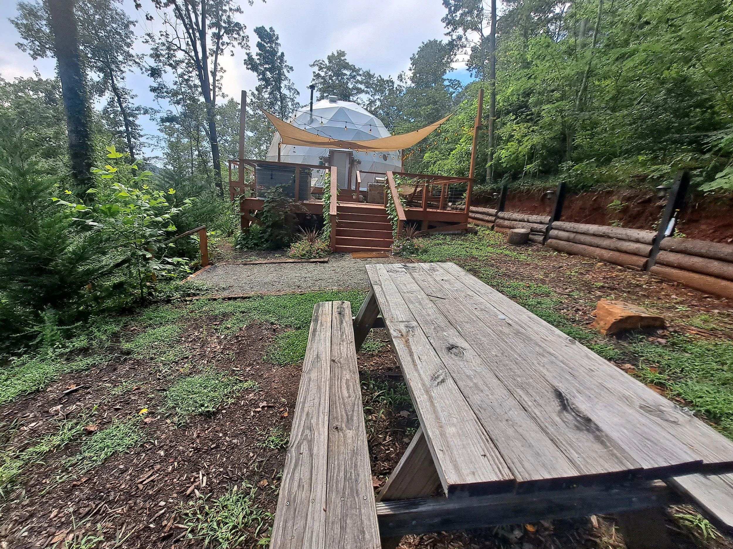 A wooden picnic table in a forest clearing with a deck in the background, featuring a geodesic dome and a shade sail.