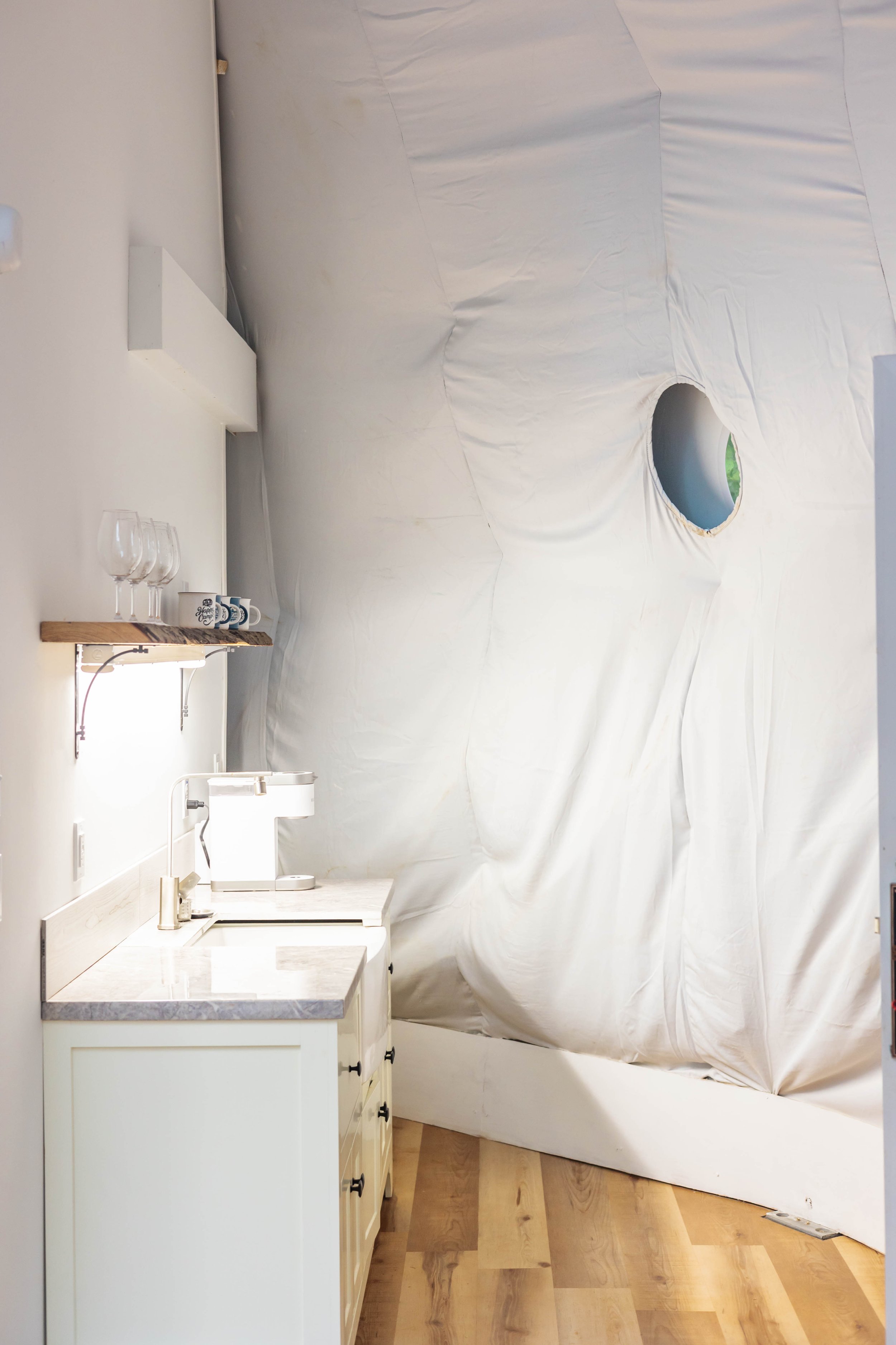 A room with a white kitchen counter, a coffee machine, wine glasses, and a shelf with mugs. The room has a large white fabric wall or ceiling with a circular hole.