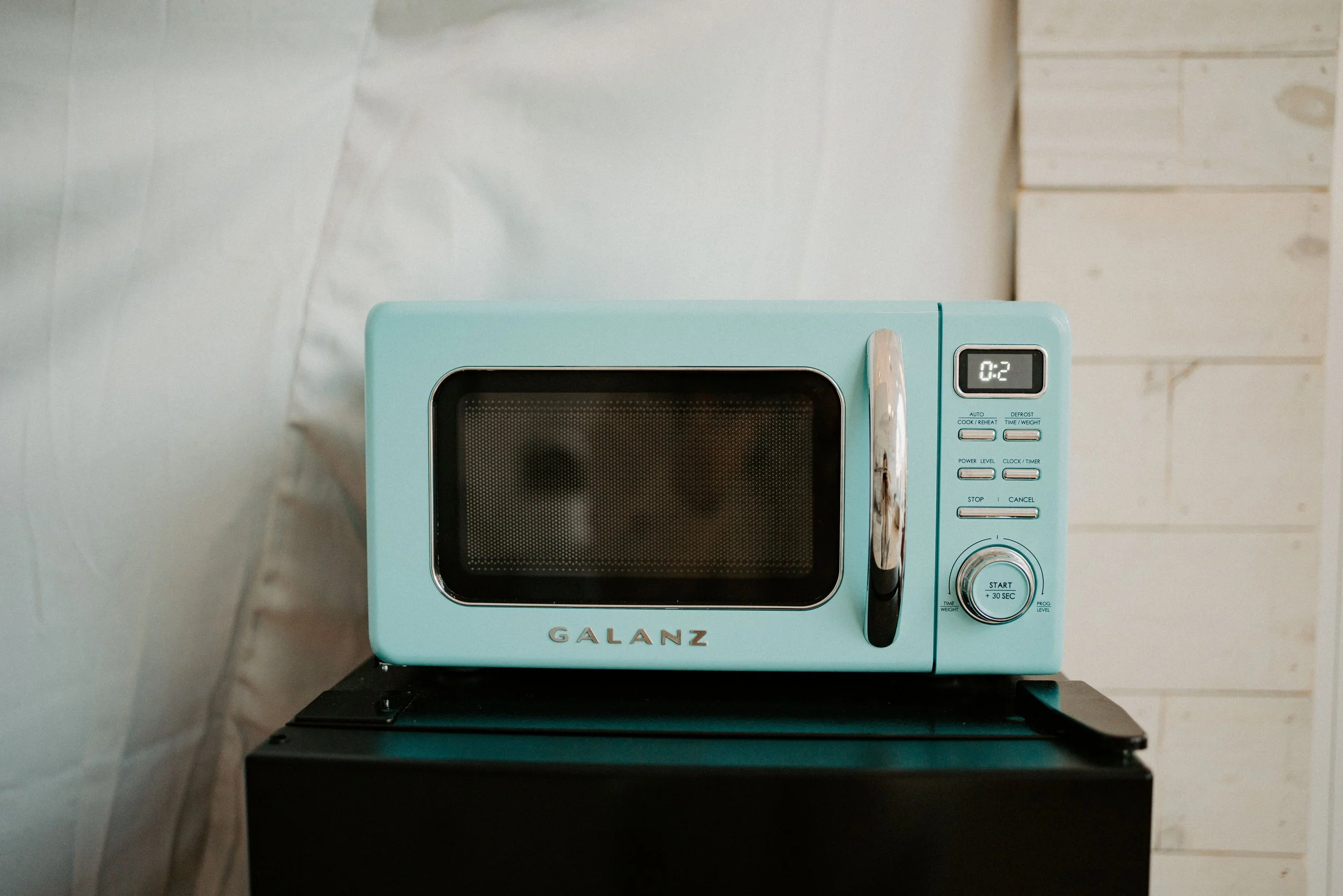 A light blue microwave oven with a black door and silver handle on top of a black surface, against a white textured wall.