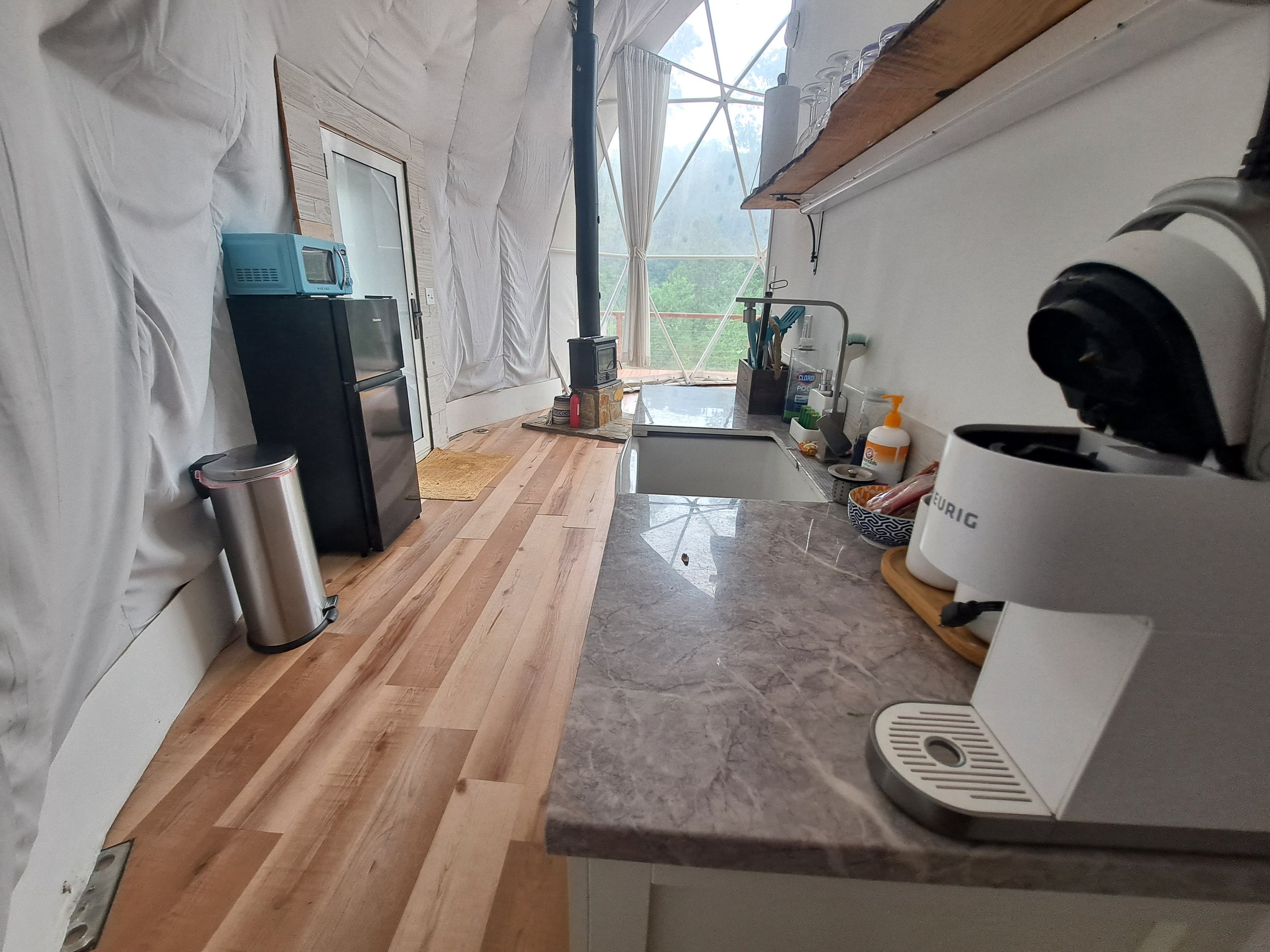 A small rustic kitchen with a marble countertop, coffee maker, drying dish rack, and utensils; a window at the end reveals a view of trees and sky, with wood flooring and a small black mini fridge with a toaster oven on top.