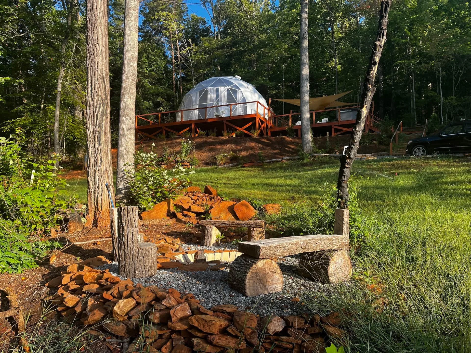 A geodesic domed structure on a wooden deck surrounded by trees in a forest, with a grassy yard and a black vehicle parked nearby.