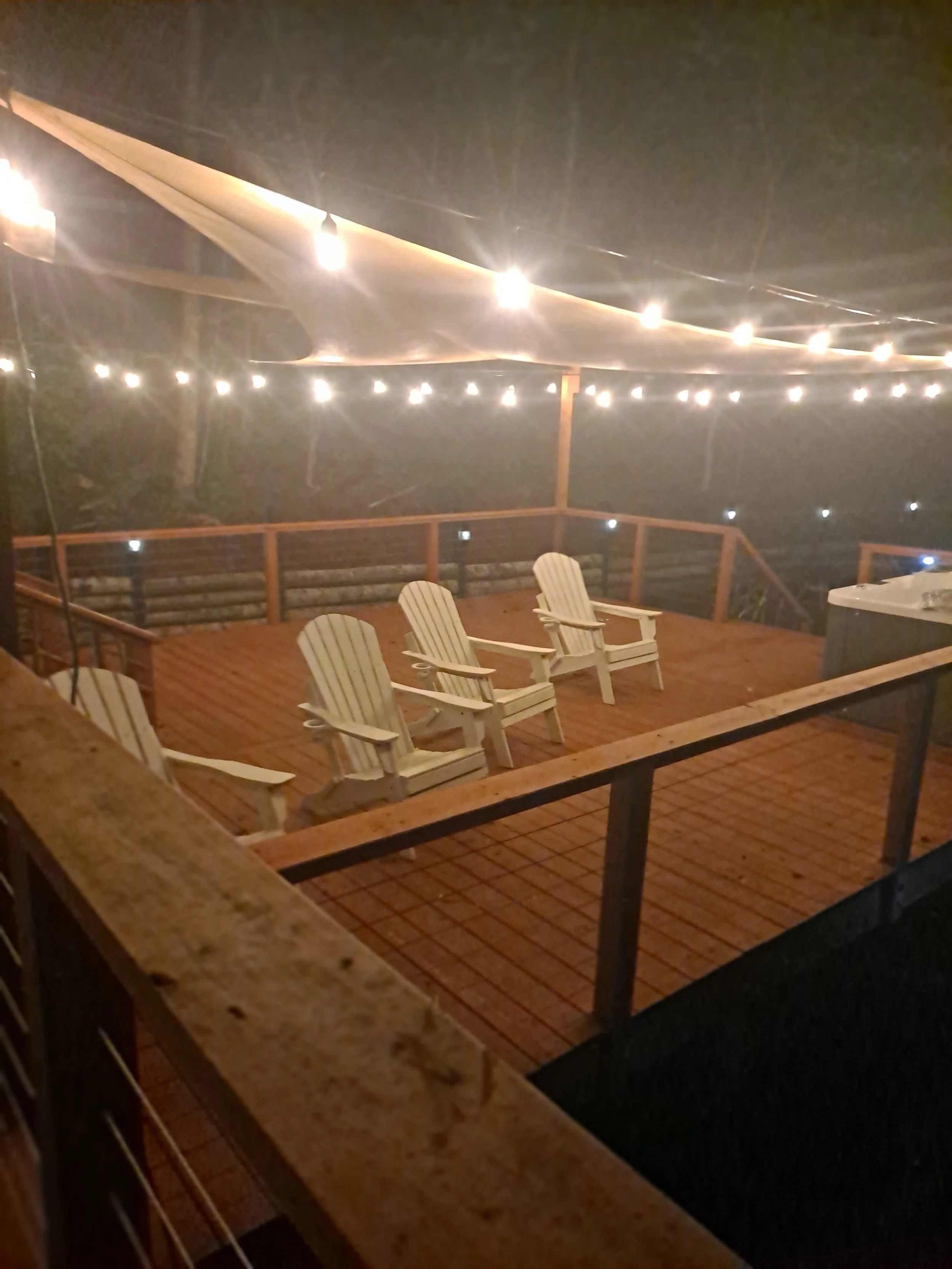 Nighttime outdoor deck with string lights, three white Adirondack chairs, and a hot tub.