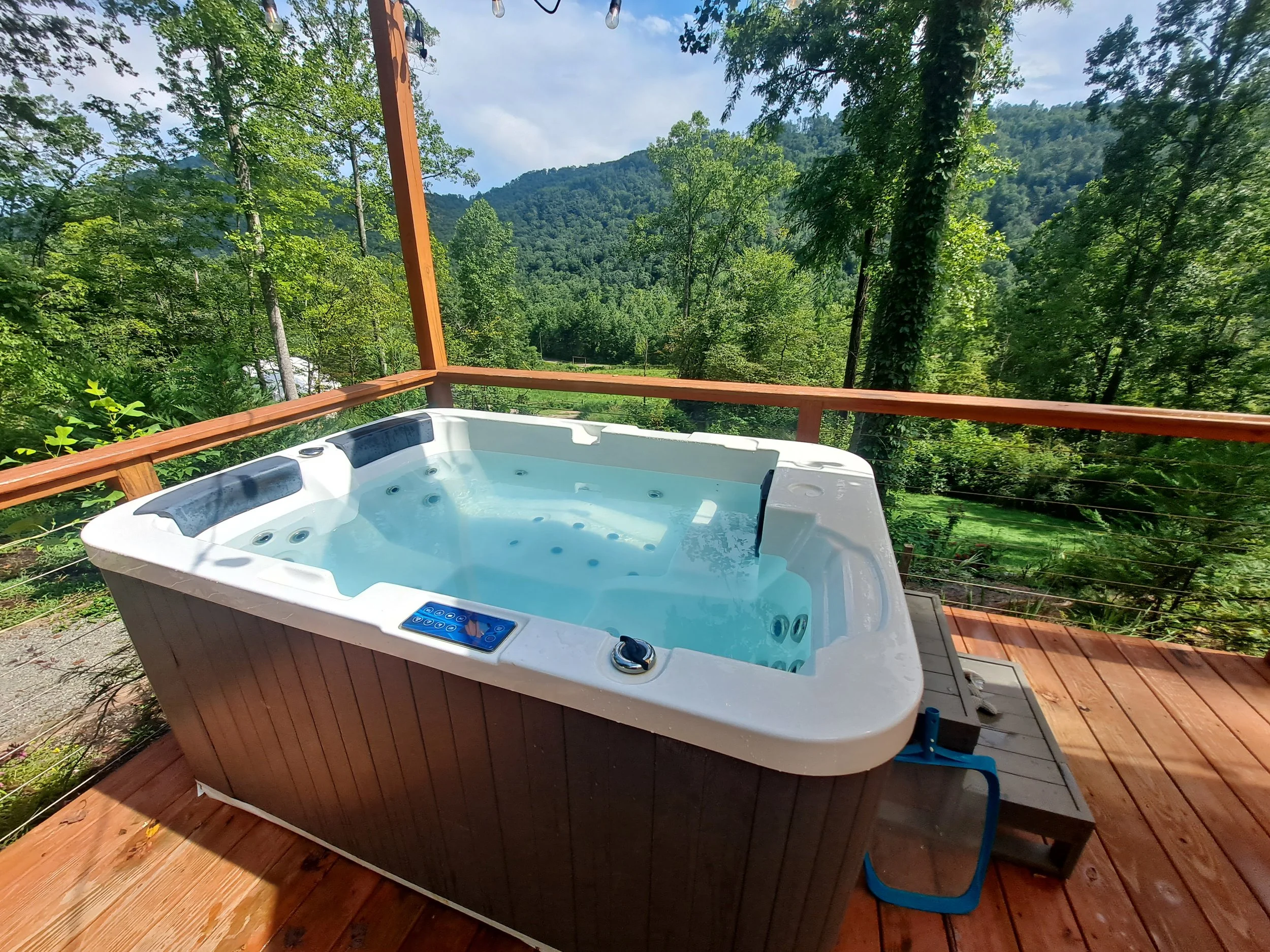 Hot tub on a wooden deck overlooking a lush green forest and mountains.