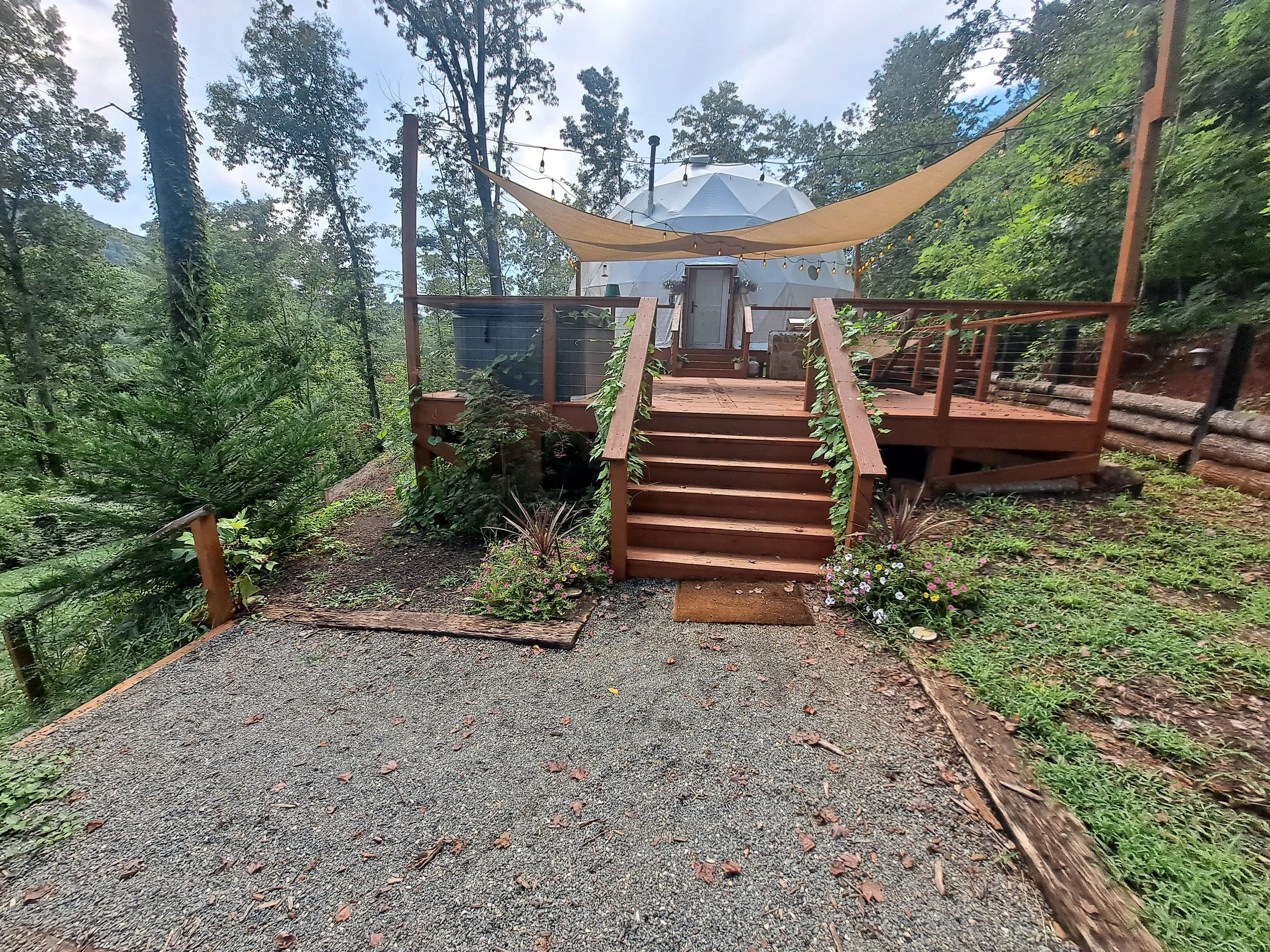 A wooden deck with stairs, surrounded by trees and plants, with a white dome-shaped tent in the background, shaded by a beige sail and string lights.