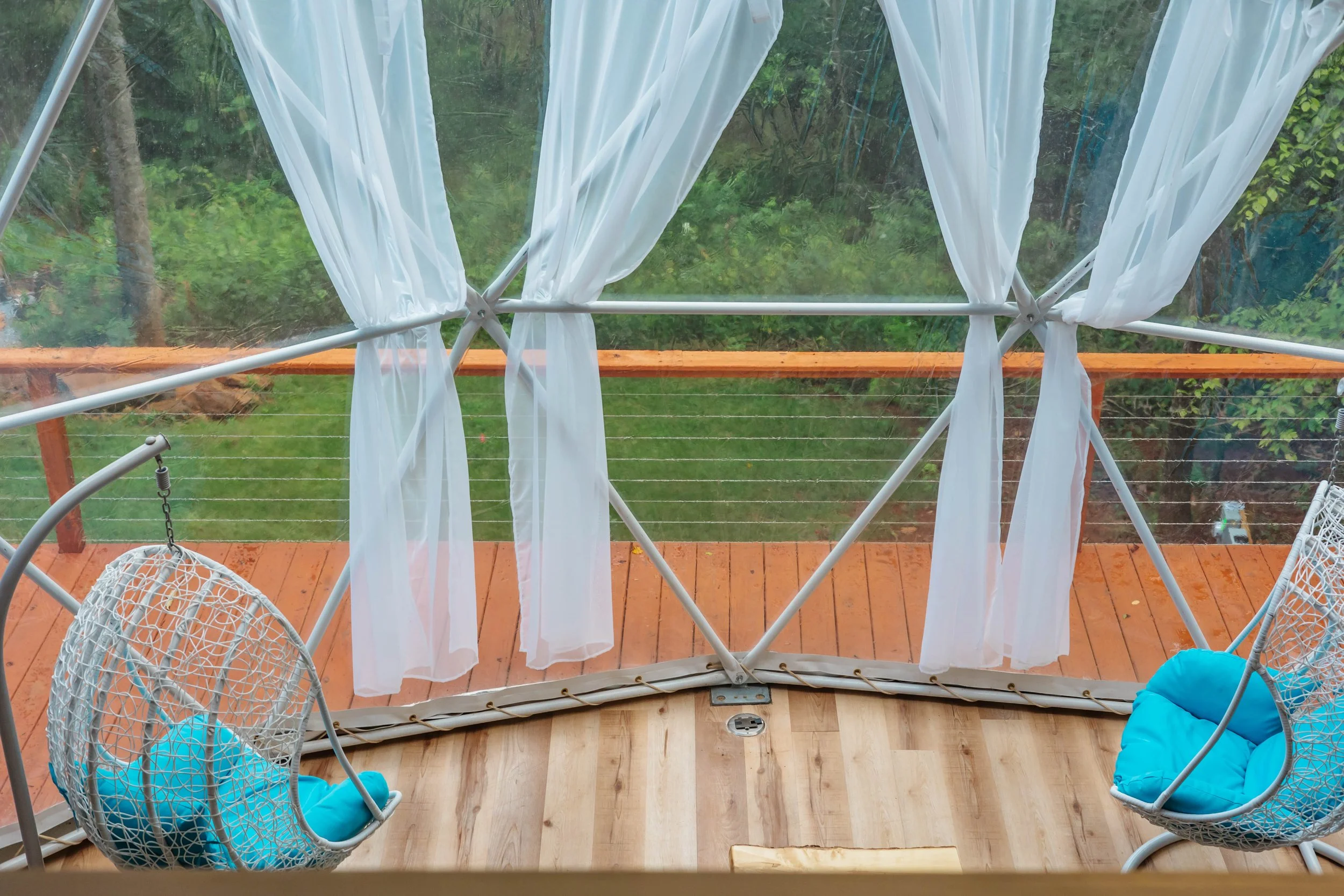 View from inside a screened porch with white sheer curtains and two hanging wicker chairs with blue cushions, overlooking a wooden deck and a green backyard with trees.