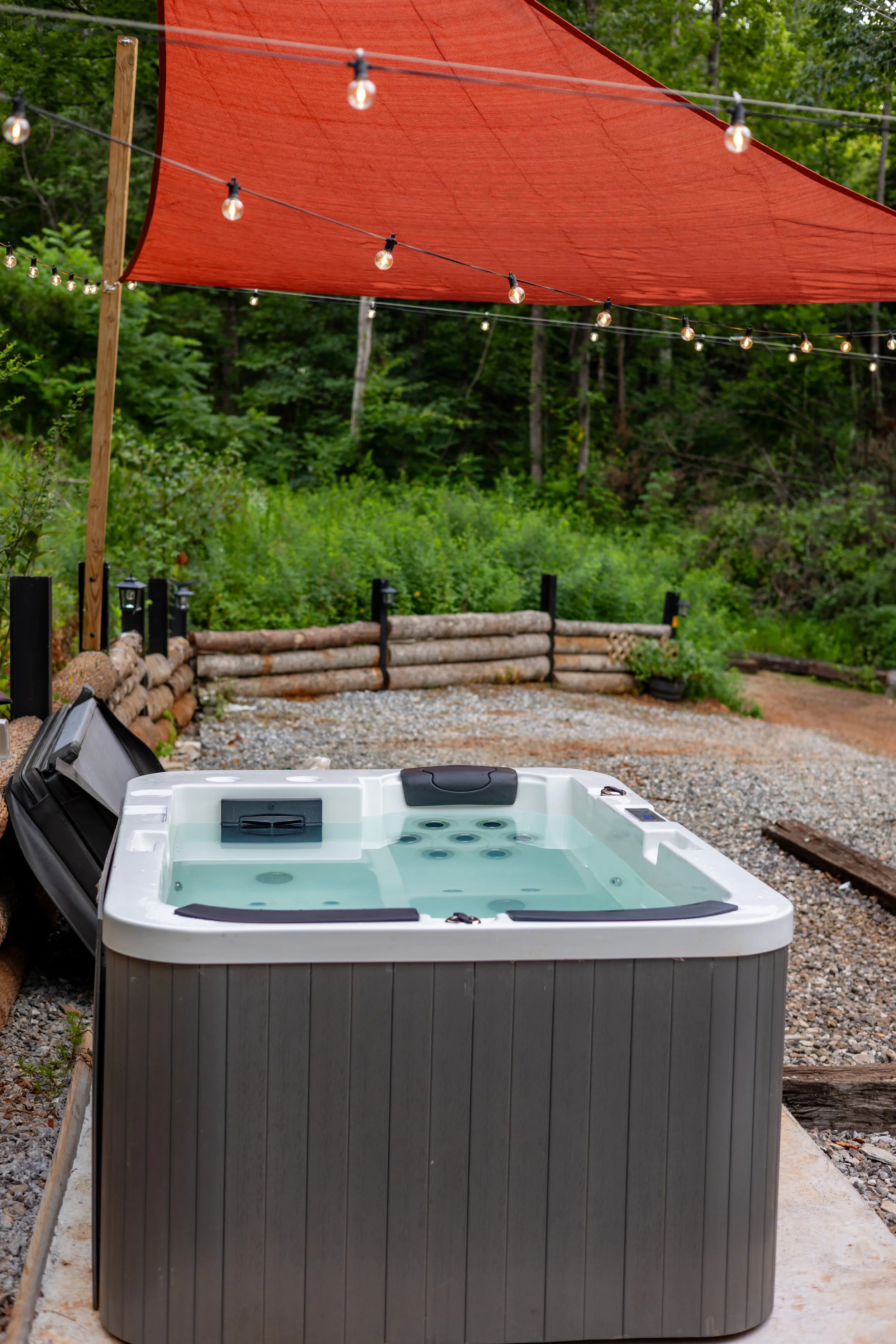An outdoor hot tub with a cover open, situated on a gravel surface under a red shade sail with string lights, surrounded by trees and greenery.
