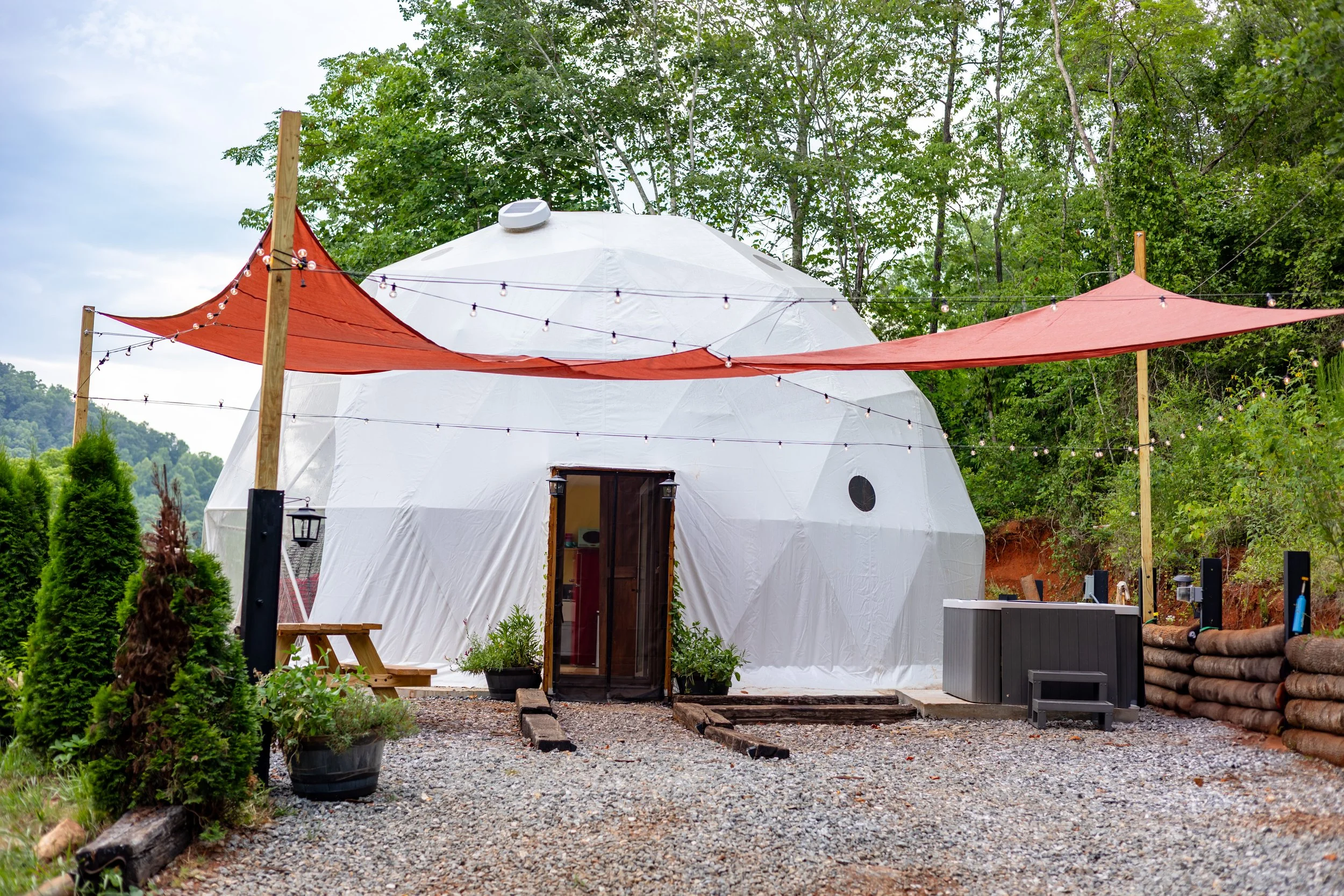 Outdoor scene featuring a large white geodesic dome with a door, surrounded by greenery, string lights, and shade sails.