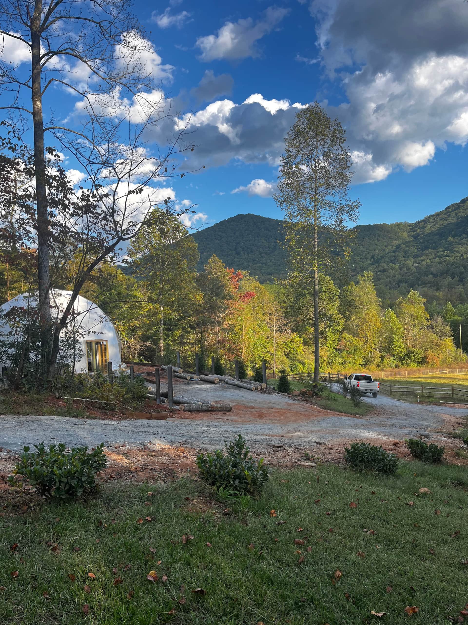 A scenic outdoor view featuring a partly cloudy sky, green trees, a mountain in the background, and a small white dome-shaped structure with a door and windows. There is a gravel driveway with logs and a white pickup truck, surrounded by natural land