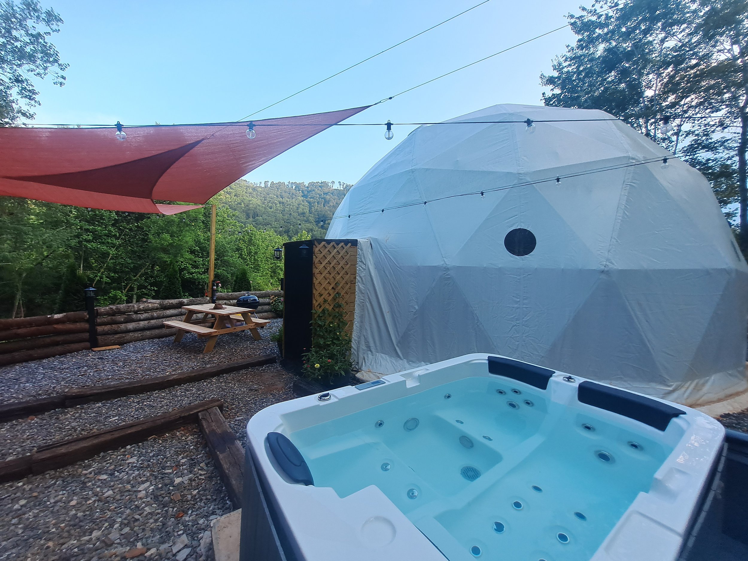 Outdoor scene with a hot tub, a white tent, a picnic table, and string lights, surrounded by trees and a hillside in the background.
