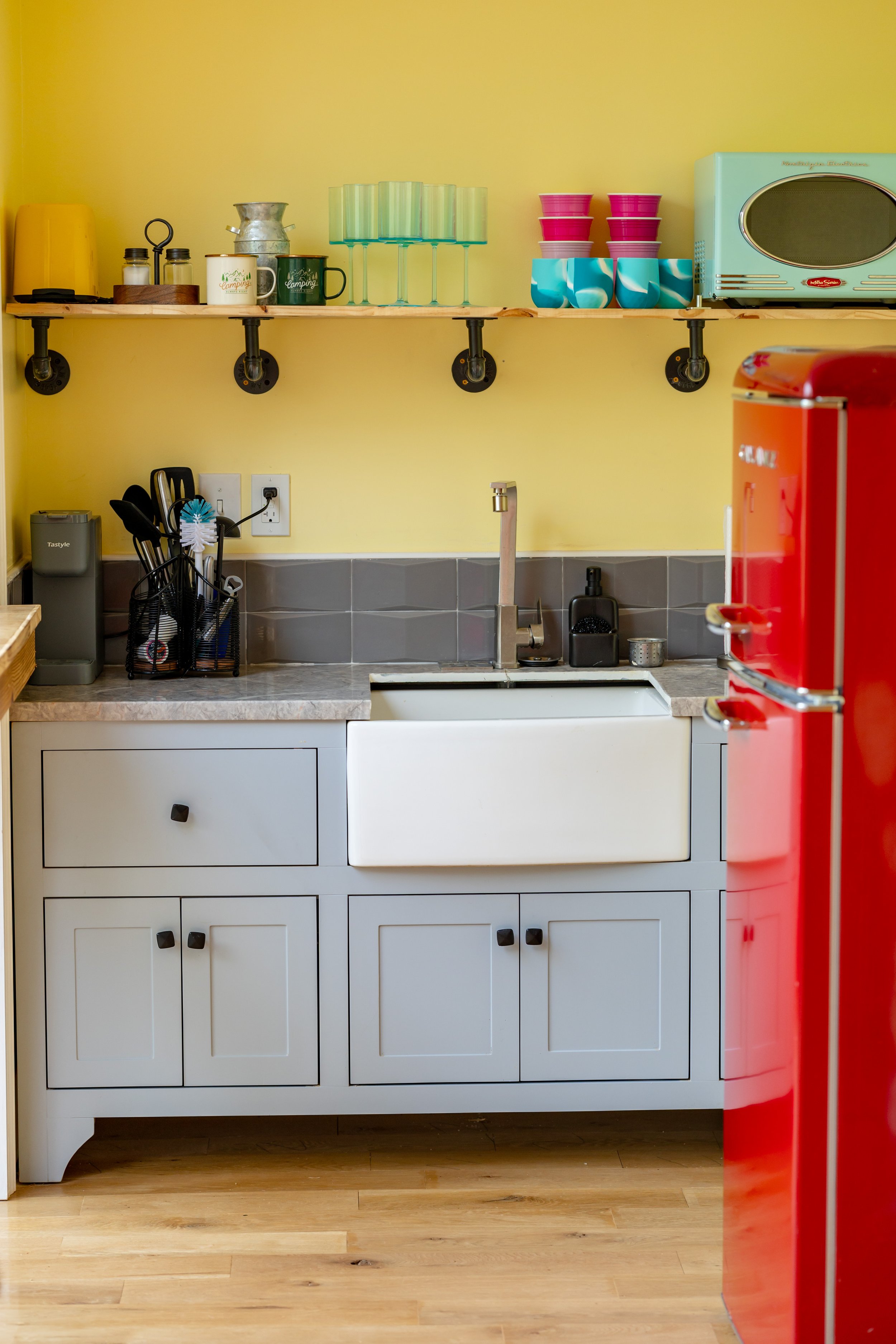 Kitchen with yellow wall, open wooden shelf holding cups, glasses, storage containers, and a toaster oven; white cabinets below sink; gray tiled backsplash; red refrigerator on the right.