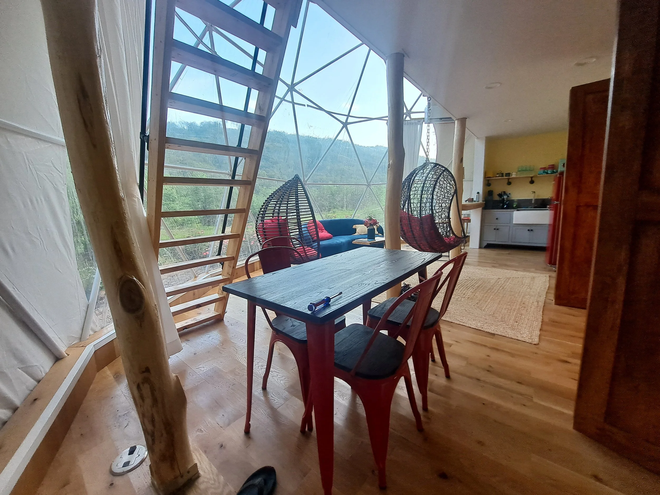 Interior of a modern geodesic dome with a wooden dining table, black chairs, hanging egg chairs, a blue sofa, and a mountain view outside.