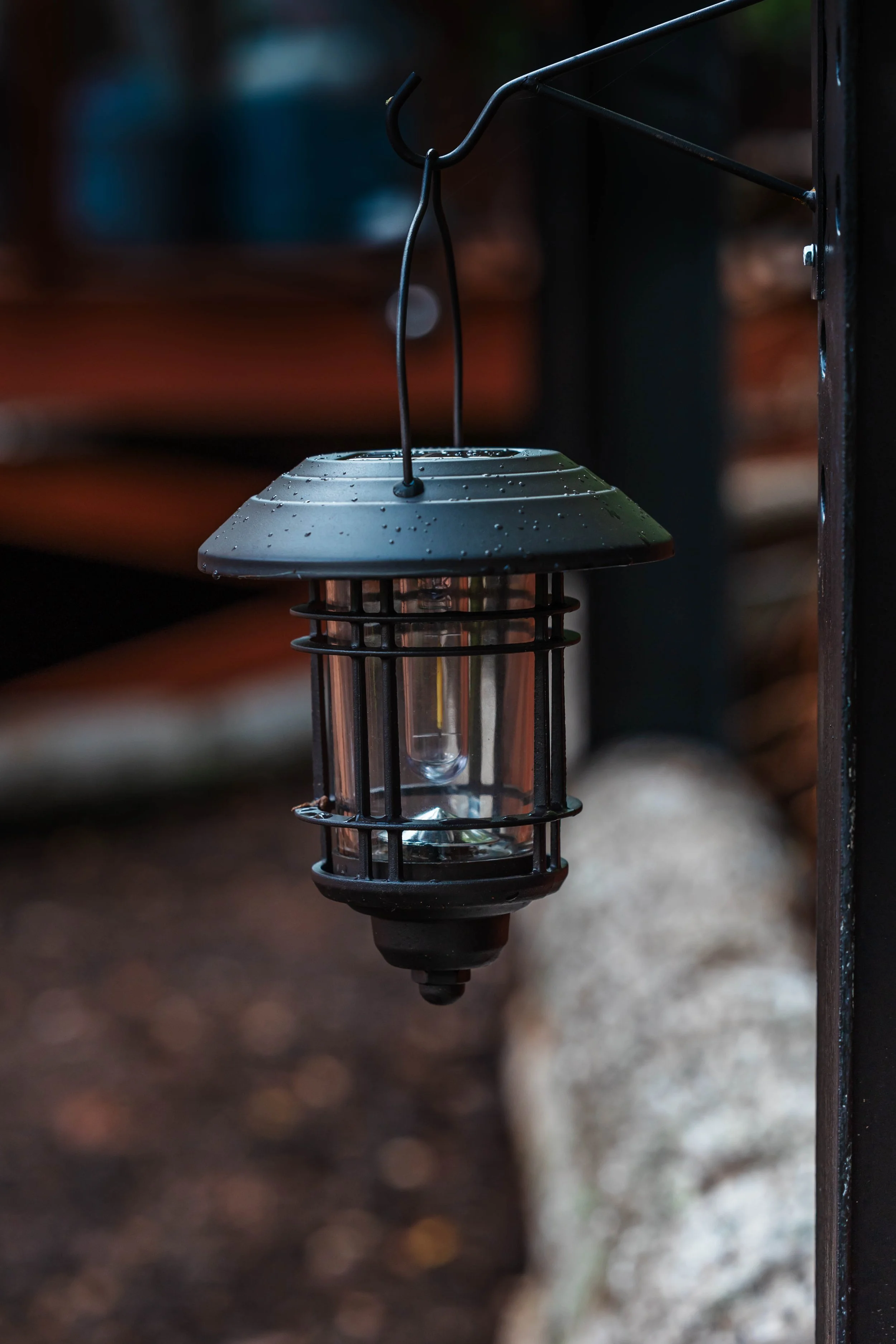 A black outdoor lantern hanging from a metal hook, with raindrops on its top, against a blurred natural background.