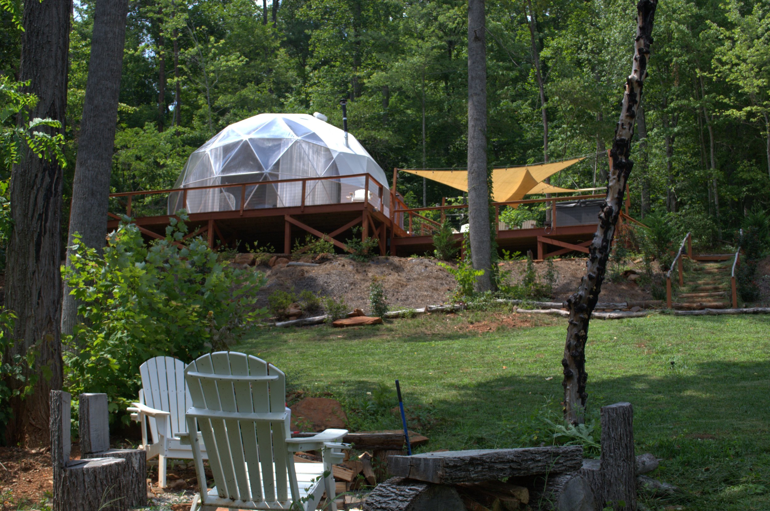 A backyard with a grassy area, several trees, and a deck with a geodesic dome structure and a yellow shade sail. Two white Adirondack chairs are in the foreground.