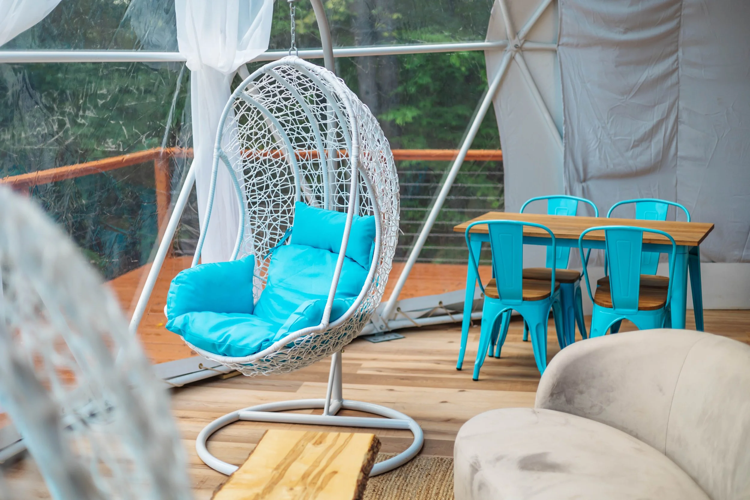 Interior of a cozy screened-in porch with a hanging egg chair with bright blue cushions, a wooden dining table with four blue metal chairs, and a beige upholstered sofa on a wooden floor, surrounded by greenery outside.