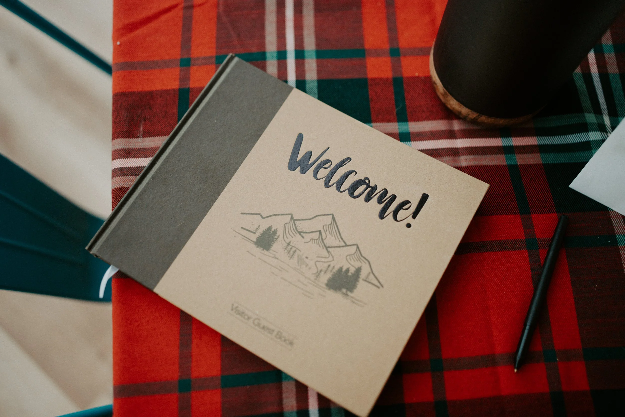 A welcome sign with a mountain illustration on a table with a red plaid tablecloth, a black pen, and a coffee mug.