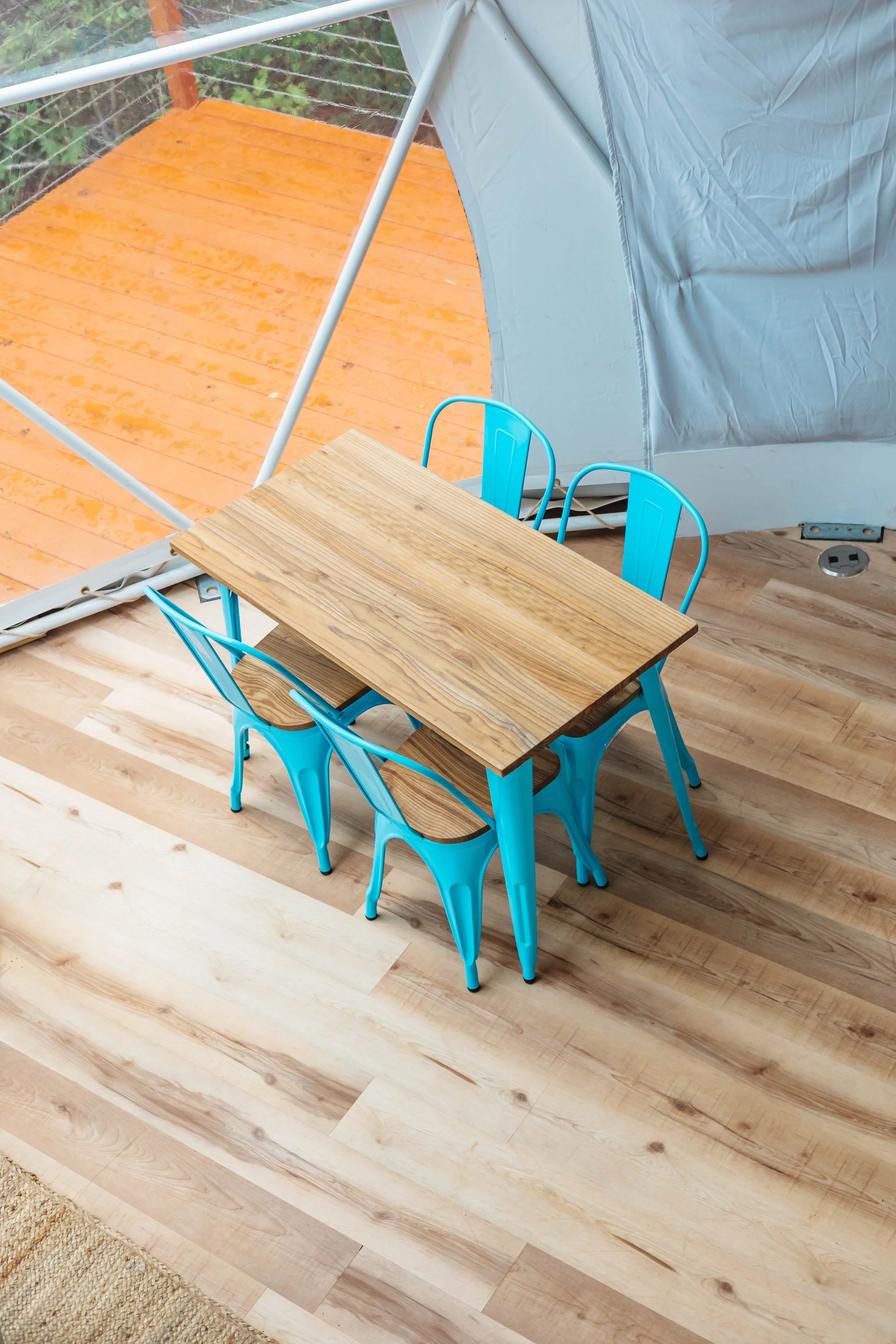 A small wooden table with four blue metal chairs is inside a screened porch with light wood flooring. The porch opens to a wooden deck outside.