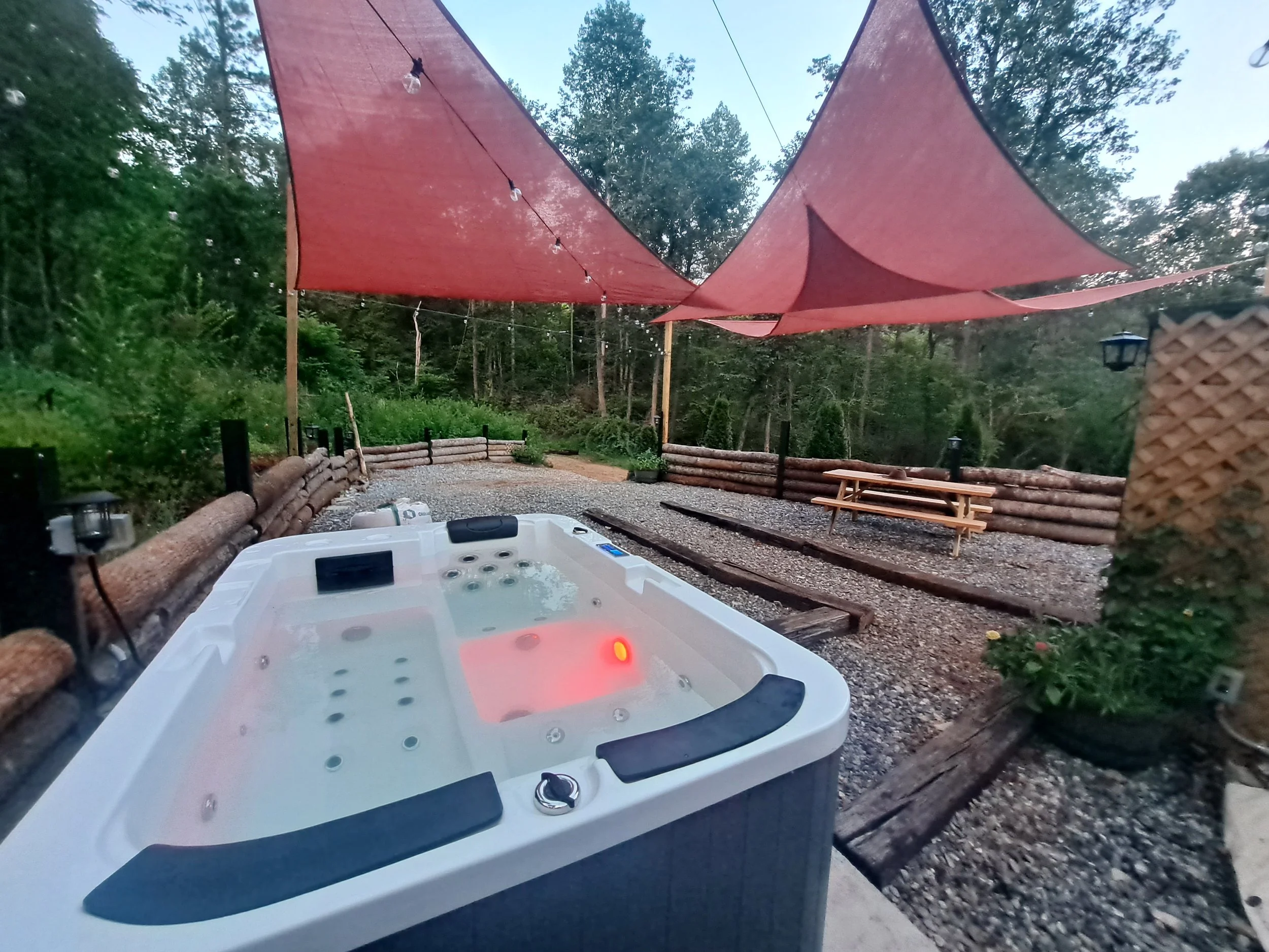 Outdoor hot tub with jets, covered by orange shade sails, surrounded by a natural woodland setting with gravel ground, wooden logs forming a low fence, picnic table, and string lights overhead.