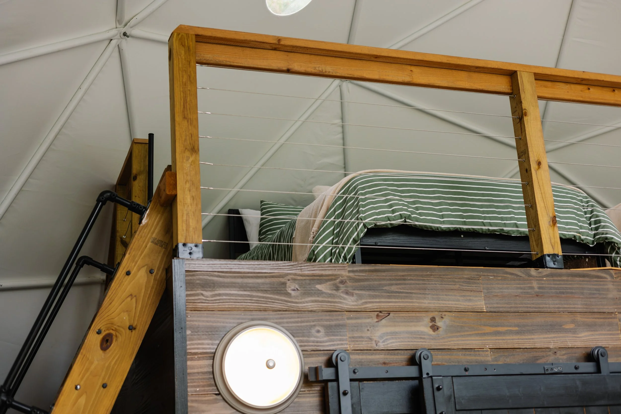 Interior of a rustic bedroom with a loft bed made of wood, featuring striped green and white bedding and pillows, inside a tent-like structure with white fabric walls and a small window overhead.