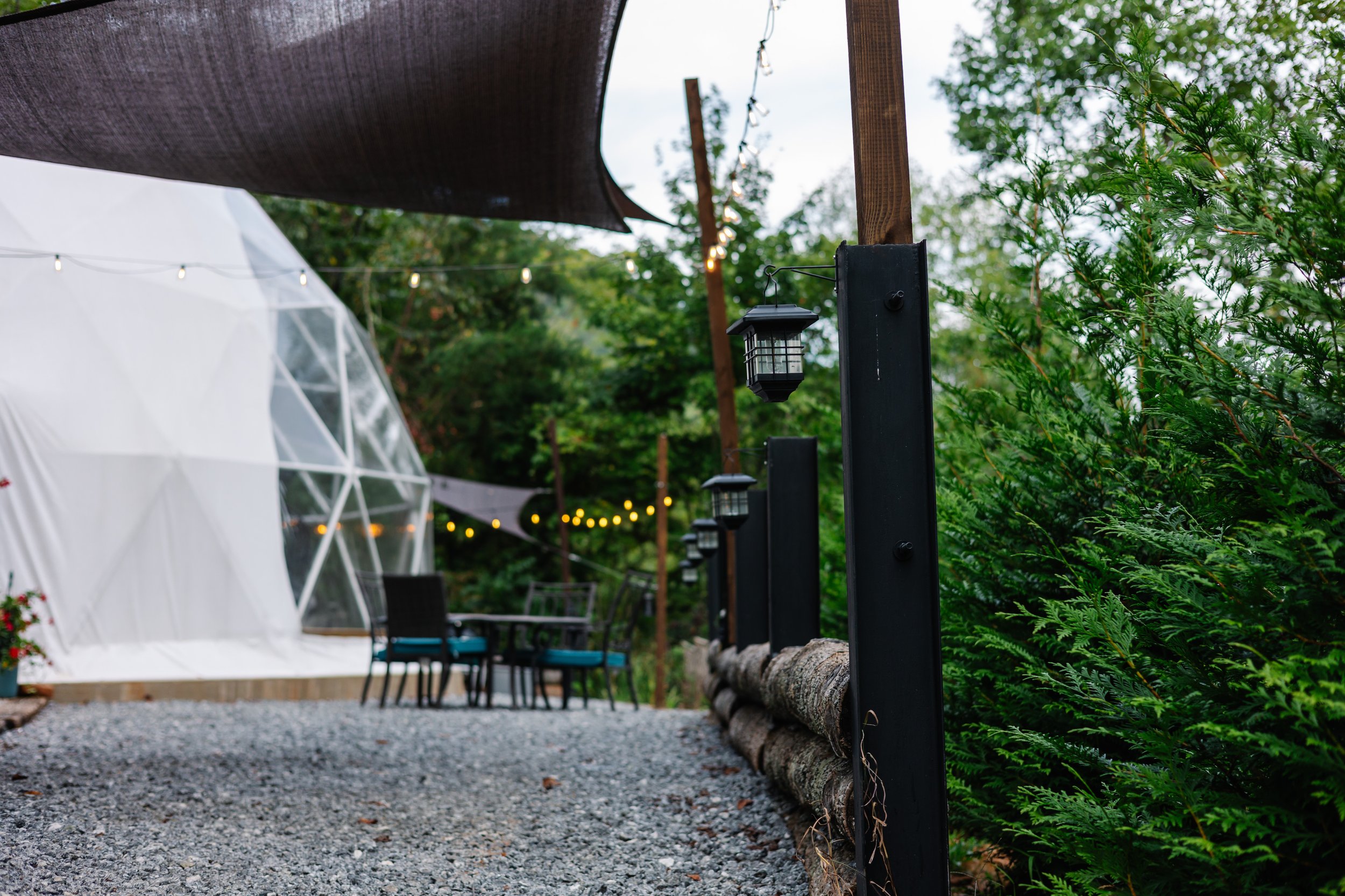 Outdoor patio with gravel ground, string lights, black lampposts, a white geodesic dome, patio table with chairs, and lush green trees.