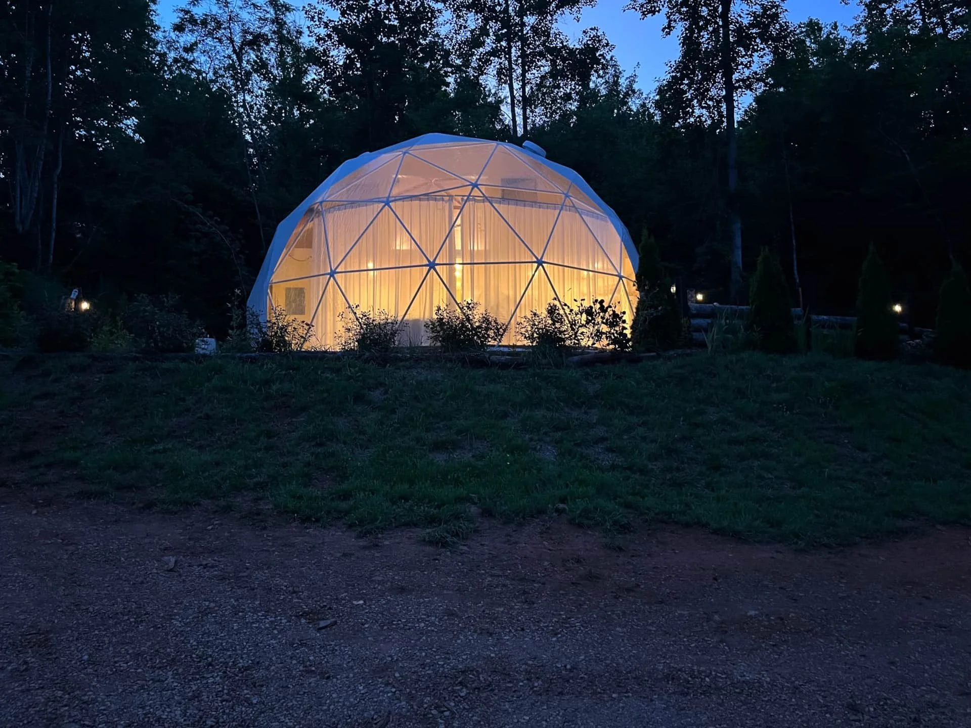 A geodesic dome tent with a warm interior light, set up in a wooded outdoor area at dusk.