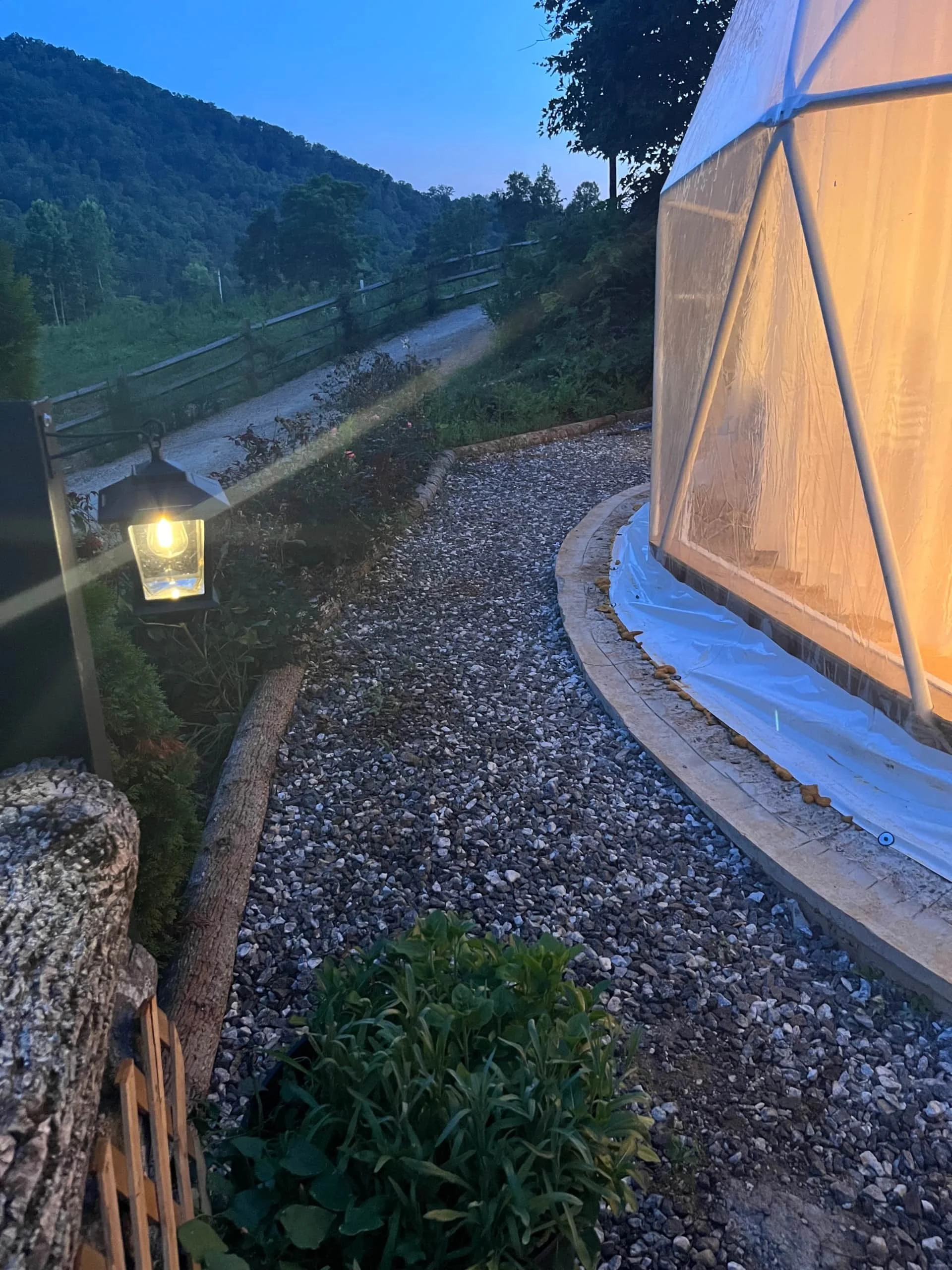 A gravel garden path outlining a yellow, dome-shaped outdoor tent with white plastic sheeting around its base, near a landscape with trees and mountains at dusk or dawn, illuminated by a single lantern-style light.