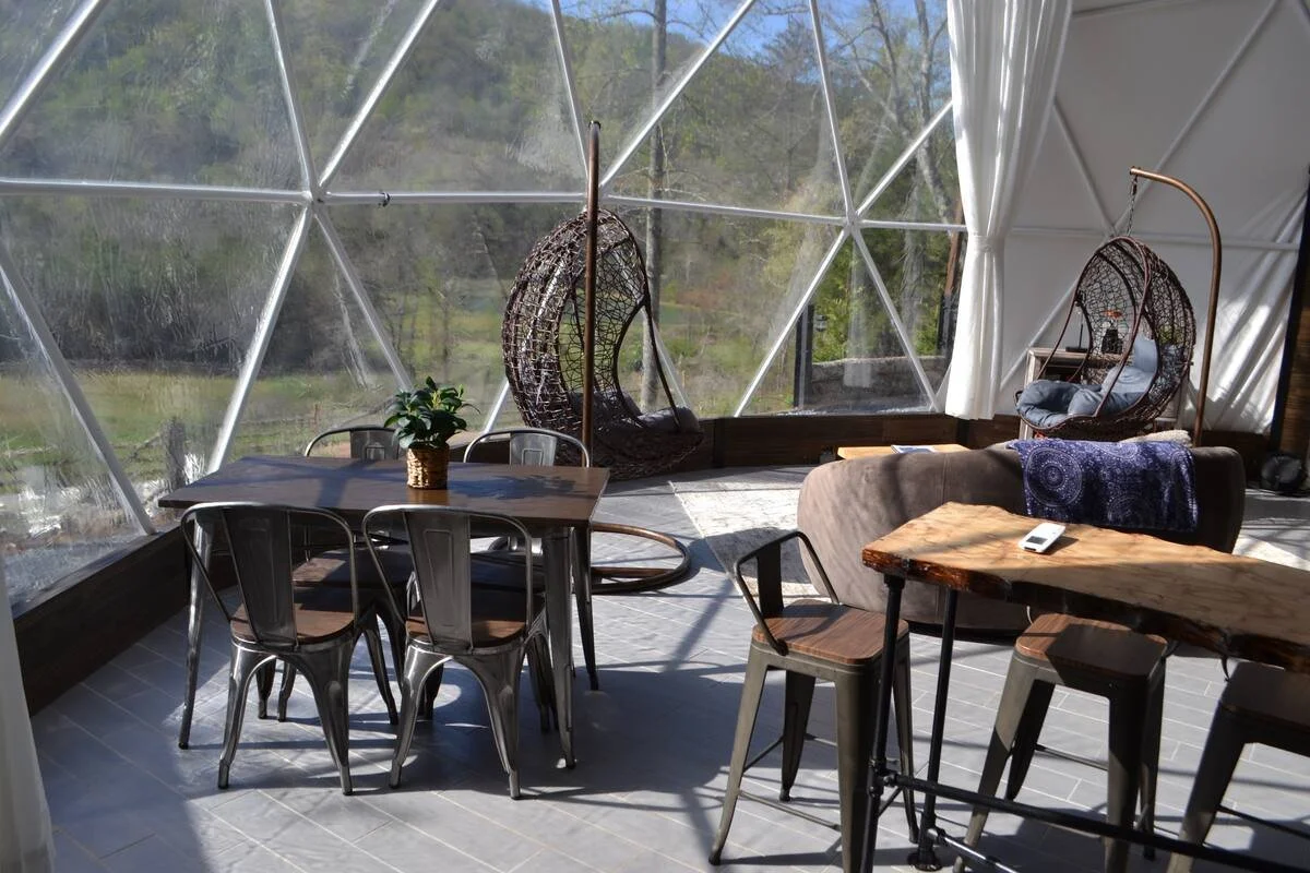 Interior of a geodesic dome with a wooden dining table surrounded by metal chairs, a sofa with a purple blanket, hanging wicker egg chairs near large windows, and a view of trees outside.