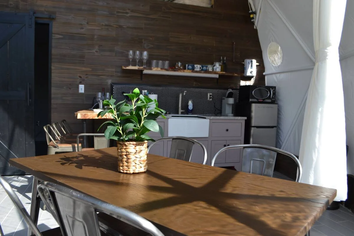 A wooden dining table with a potted plant in a woven basket on it, surrounded by metal chairs, in a space with a kitchenette and natural light.