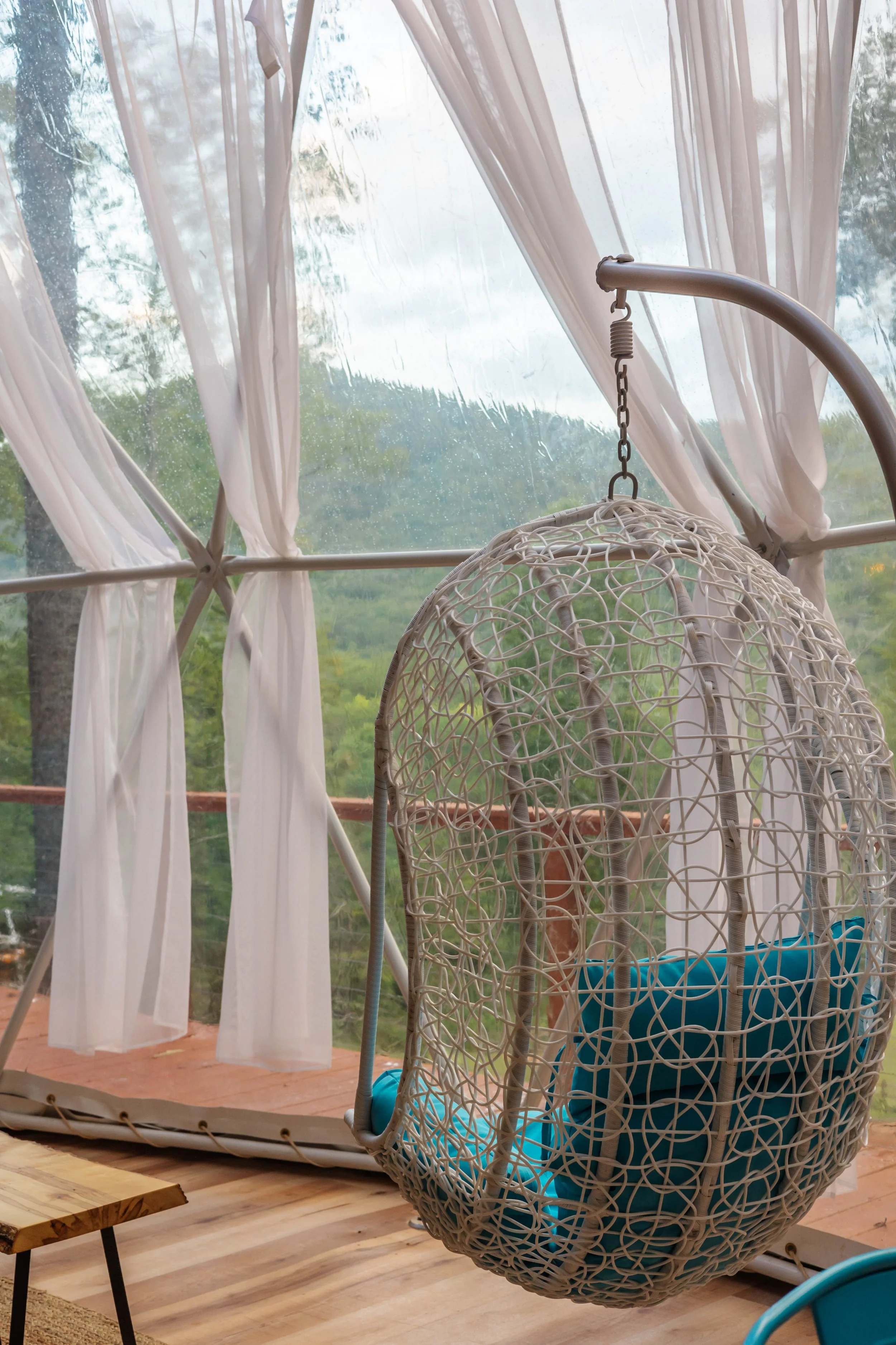 A hanging wicker chair with blue cushions in front of a screened porch with white curtains and view of trees and mountains outside.
