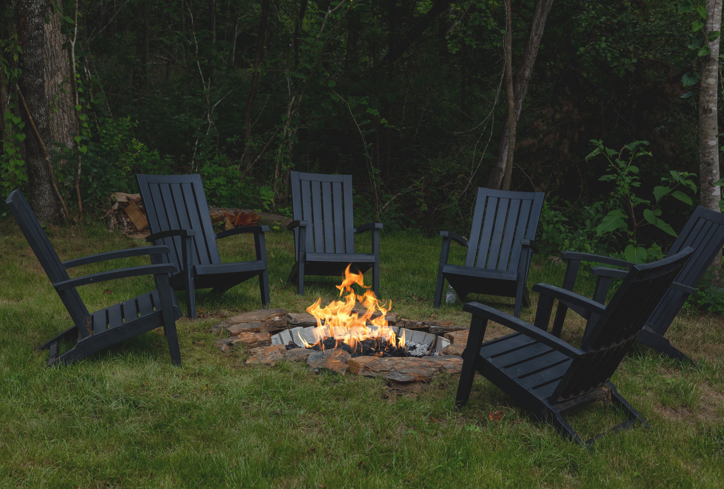 Six dark blue Adirondack chairs arranged in a circle around a campfire on a grassy patch near trees.