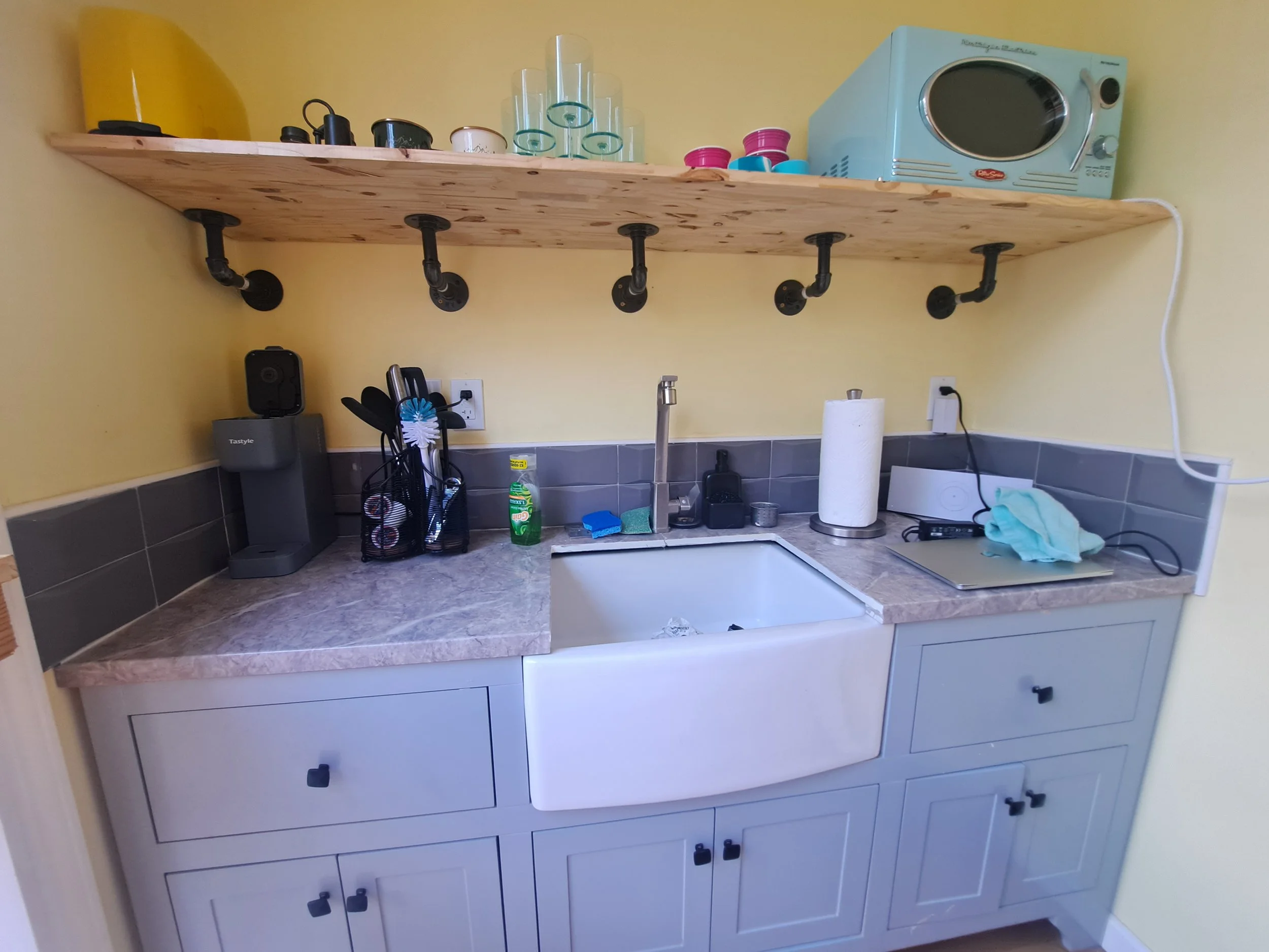 Small kitchen corner with gray cabinets, a farmhouse sink, a gray marble countertop, a wooden shelf with black pipe supports, various small appliances, cleaning supplies, a paper towel roll, a soap dispenser, and a microwave oven.