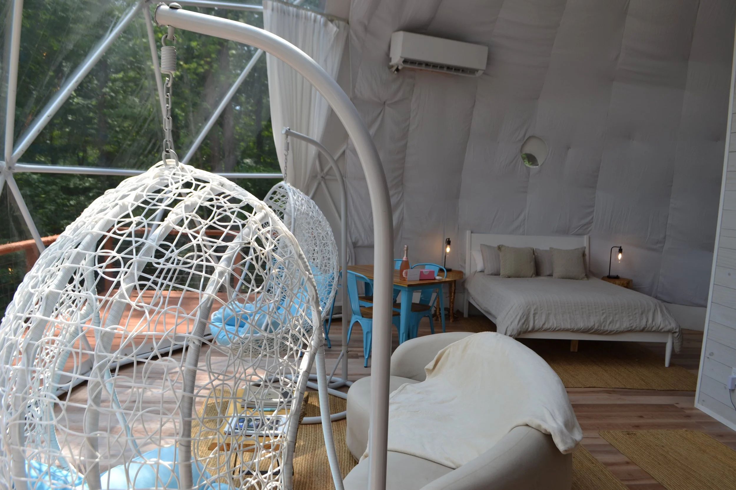 Interior of a cozy tent with a bed, nightstands, a small desk with chairs, hanging chairs, and a window showing greenery outside.