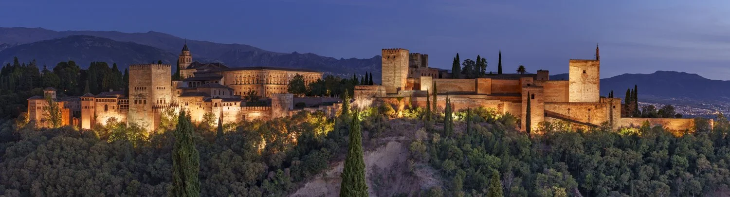 Night view of the Alhambra Palace and fortress illuminated, with mountains in the background and a dark blue sky.