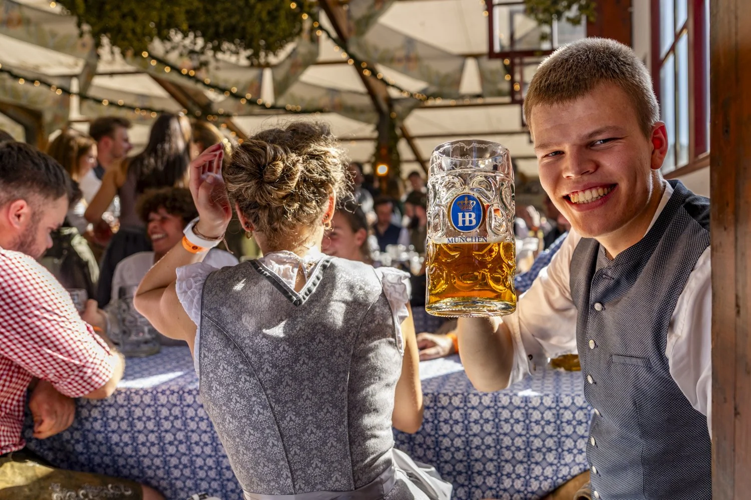 A young man smiling and holding a large mug of beer at a festive gathering, with people in traditional clothing around him at an indoor event with string lights and a decorated wooden ceiling.
