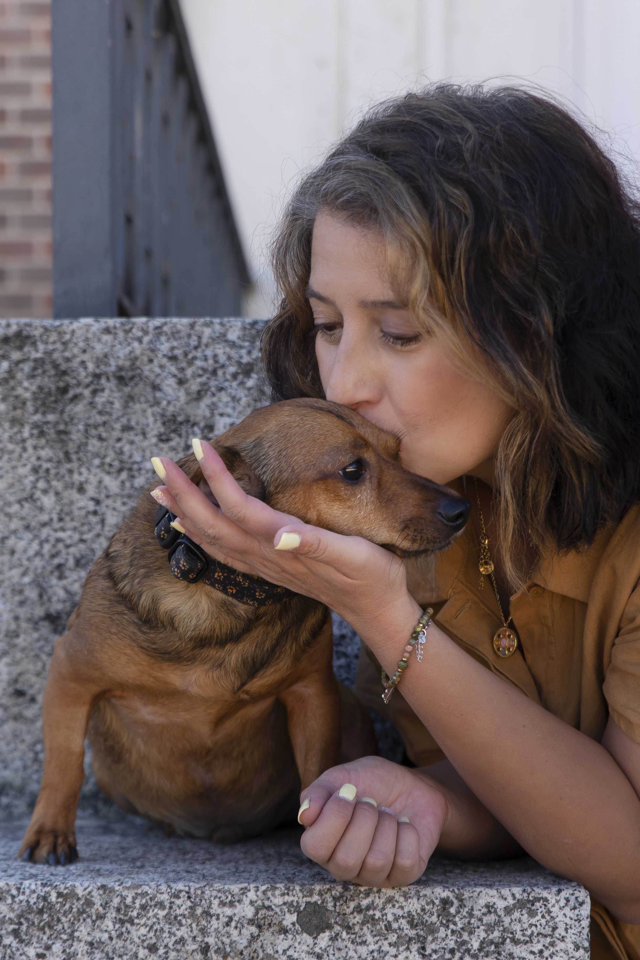 Mujer besando a un perro pequeño marrón en la cabeza, mientras lo sostiene con una mano. Están sentados en un banco de piedra. La mujer lleva una pulsera, collar y ropa de color marrón claro.