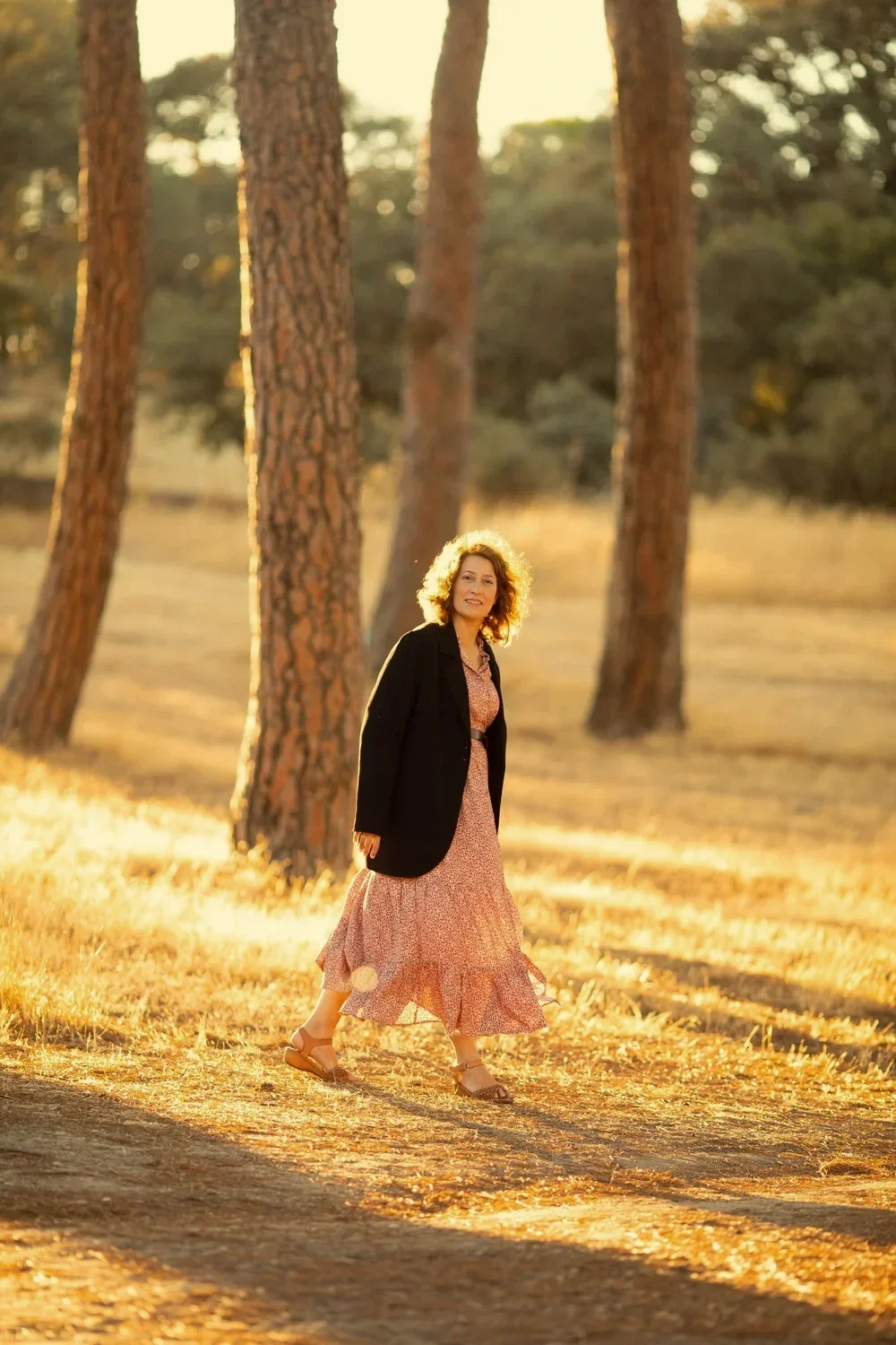 Mujer caminando por un bosque con árboles altos, vestida con un vestido largo y un saco negro, durante el atardecer con luz dorada.