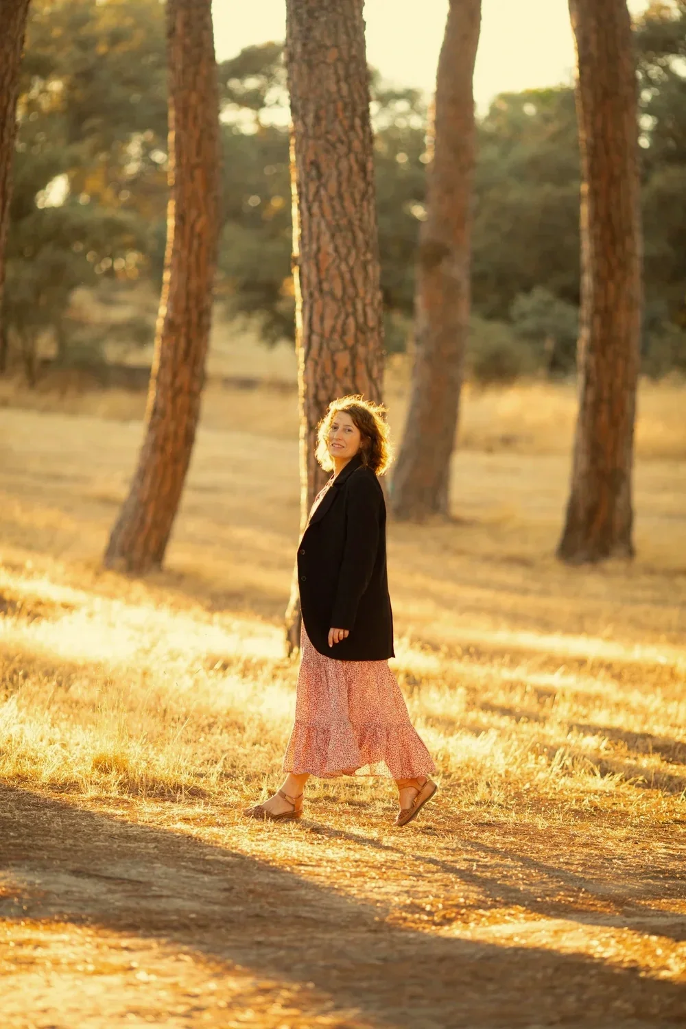 Mujer caminando entre árboles en un paisaje al atardecer, vestida con falda larga y chaqueta negra.