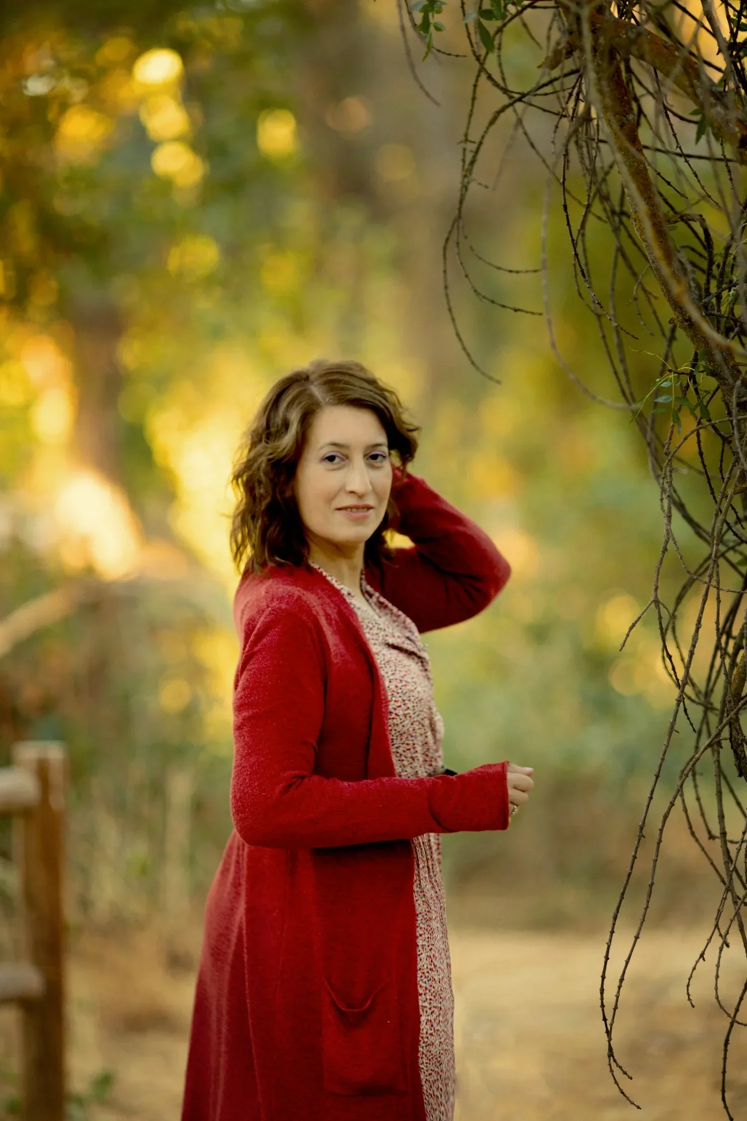 Mujer con cabello rizado de cabello marrón oscuro, usando un abrigo rojo y vestido de patrones claros, en un entorno natural con árboles y luz cálida en el fondo.