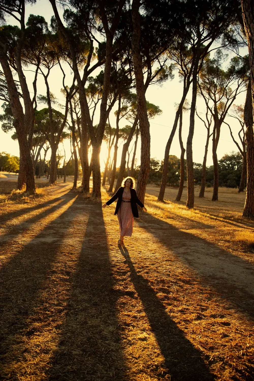 Persona caminando entre árboles durante la puesta del sol, con largos sombras proyectadas en el suelo.